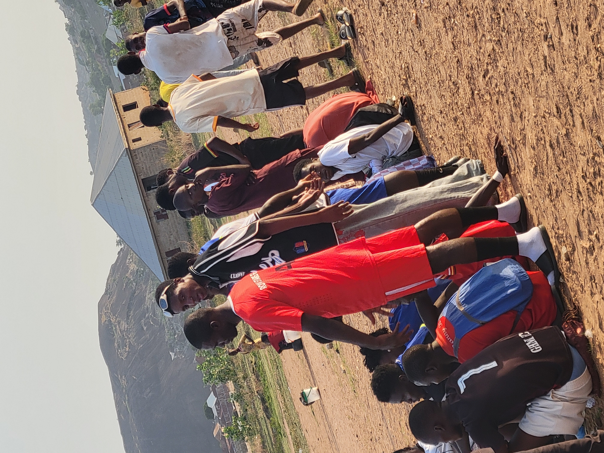 A wide-angle, eye-level shot depicts a group of young men in Ibo, Nigeria, engaged in an outdoor activity. The men are positioned on a reddish-brown dirt surface. They are wearing a variety of clothing, including sports jerseys, shorts, t-shirts, and socks. Some have backpacks near them. A building with a tin roof and a partially constructed brick structure is visible in the background. Further in the distance, a large, rocky mountain formation is seen against a hazy sky. The environment appears to be a somewhat arid, grassy field.