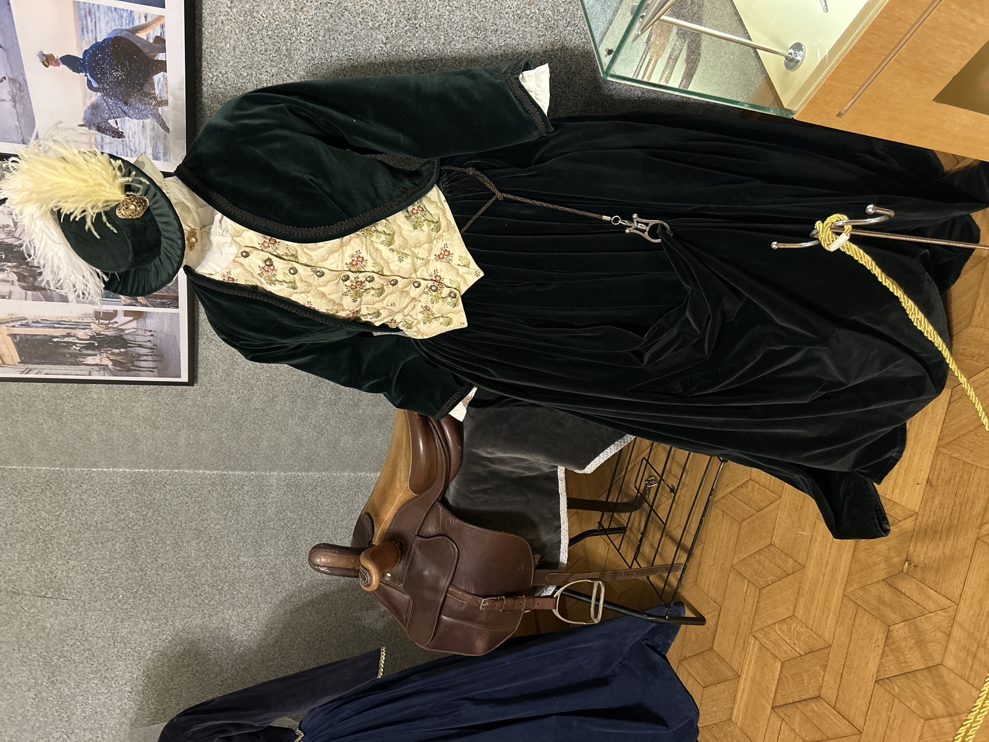 The image shows a display of historical equestrian attire in a museum. In the foreground, a mannequin is dressed in a dark green velvet jacket, a cream-colored waistcoat with a floral pattern, and a full black velvet skirt. The mannequin wears a matching dark green hat adorned with white and yellow feathers and a gold brooch. A leather saddle on a display stand is to the left of the mannequin, and a dark blue velvet dress is visible to the far left. The background features a textured grey wall with framed historical photographs of people on horseback. The floor is made of light brown wooden parquet. A glass display case with metal stands is partially visible on the right side of the image. The scene appears to be indoors, likely in a museum exhibit.