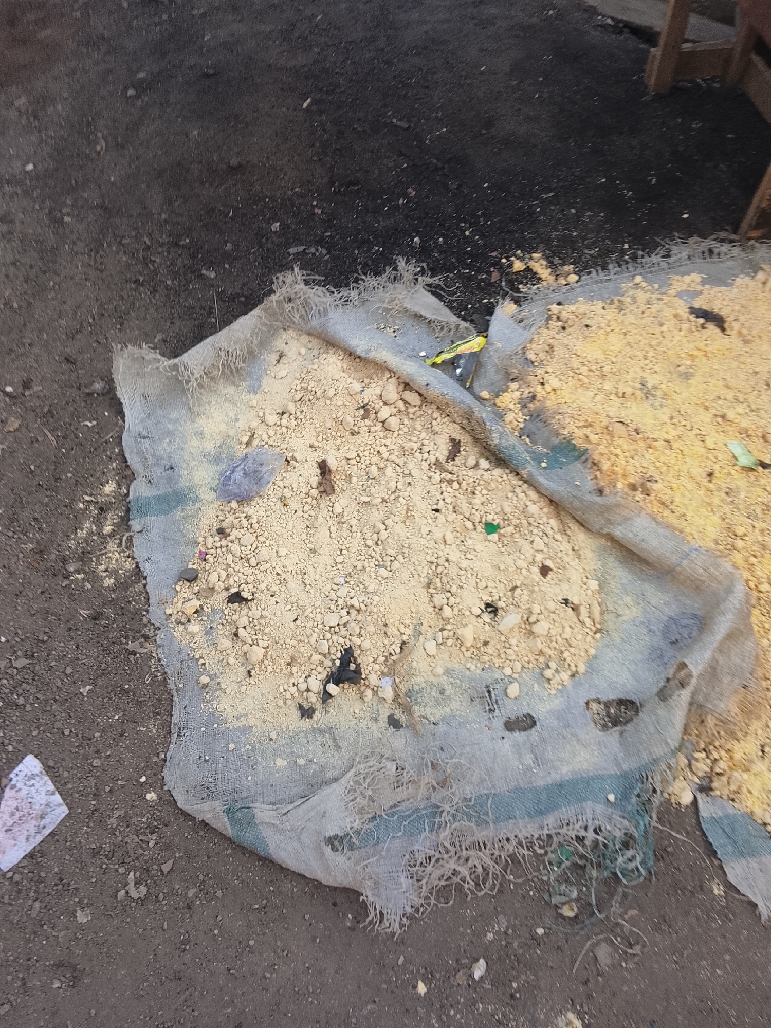The image depicts an outdoor scene, likely in a marketplace or a roadside area in Abuja, Nigeria. In the foreground, a tattered piece of burlap fabric is spread on the ground, covered with a light yellow granular substance, possibly sand or grain. There are small pebbles and debris scattered across the fabric and surrounding dirt. To the right, another similar piece of fabric with the same granular material is visible. In the background, the ground appears to be dark, coarse dirt or asphalt, with some wooden structures and possibly a portion of a table or bench to the far right. A small piece of white paper with a pattern is visible on the dirt to the lower left. The overall impression is one of a disheveled or abandoned area where goods may have been stored or processed.