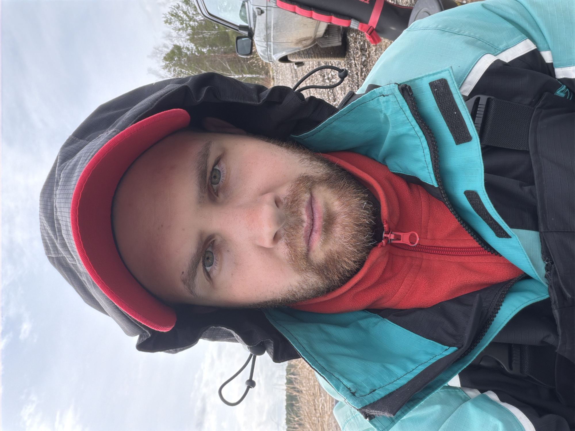 A selfie taken in an outdoor setting, possibly near Chaikovsky, Russia. A man with a beard wears a hooded jacket with a red inner layer and a blue and black outer layer. The background includes a car, a forest, and a cloudy sky. The ground appears to be covered in dry grass or vegetation.