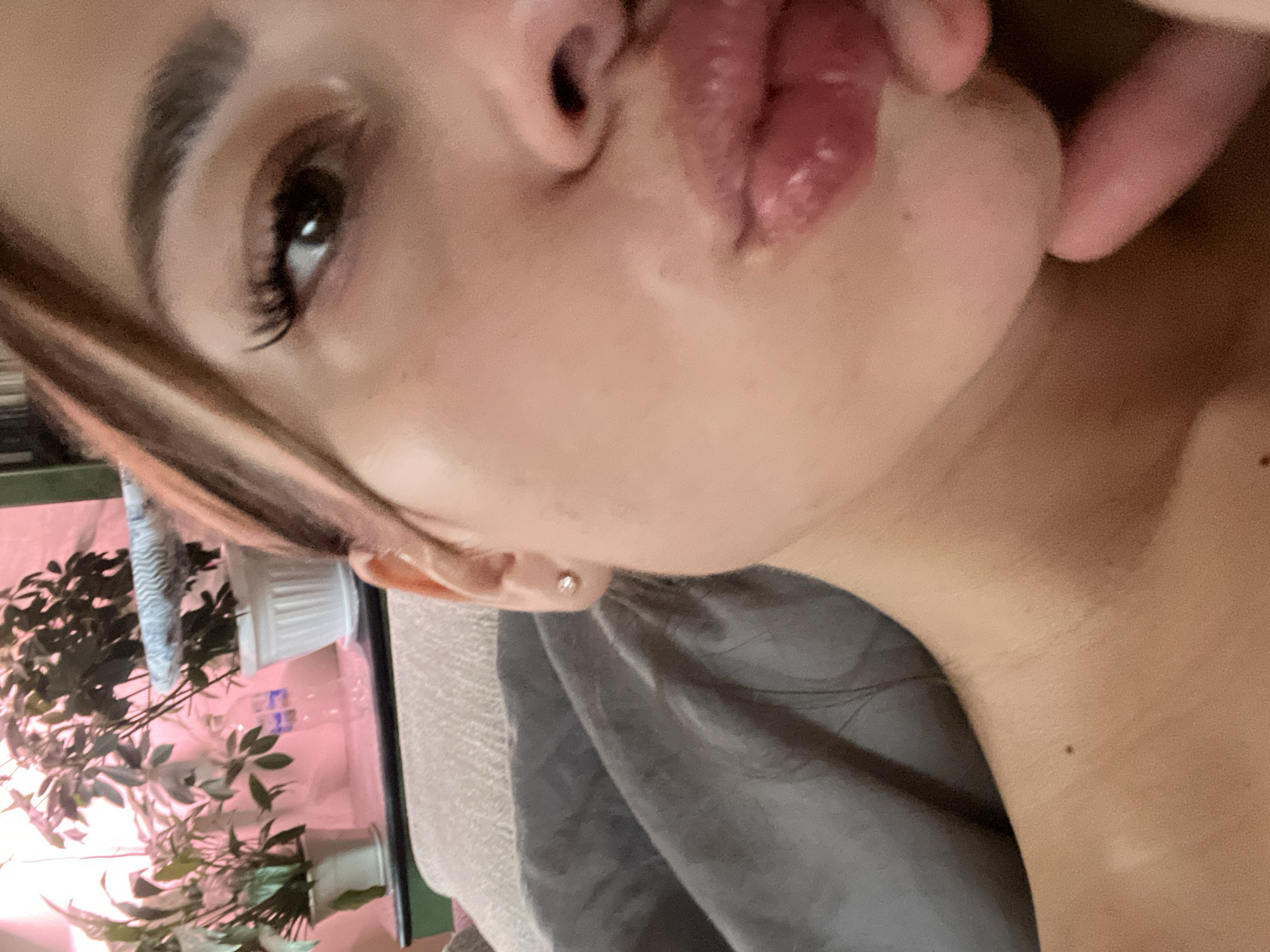 A close-up portrait of a woman with brown hair and fair skin. She has full lips and is making a kissing face. Her right eye is visible, showing long eyelashes. She is wearing an earring in her right ear. Her neck and upper chest are visible. In the background to her left, there's a potted plant with green leaves, situated on a surface illuminated with pink light. The background is indistinct. There is no evidence to suggest that the image was taken in Ust-Ilimsk, Russia.