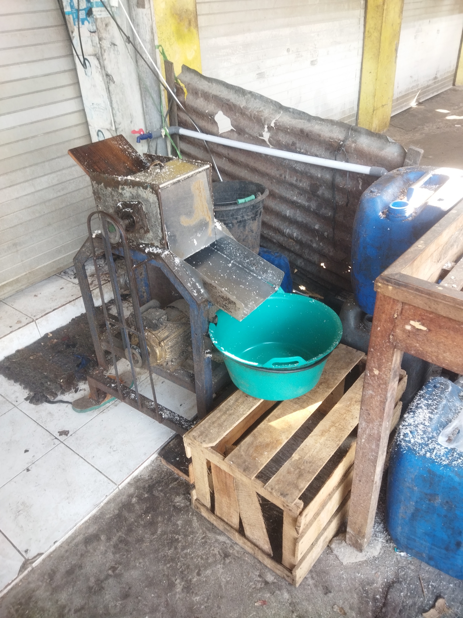 This is a medium-shot, eye-level photograph of a food processing machine with a motor and a teal-colored basin, likely in an outdoor or semi-outdoor setting in Sukoharjo, Indonesia. The machine appears to be used for grating or processing food items, as evidenced by white residue on the machine and in the basin. To the right of the machine, a wooden crate is placed on the ground, supporting the basin. Behind the machine, a blue plastic barrel is visible, along with a corrugated metal sheet acting as a wall or cover. The ground is a mix of white tiles and concrete, with debris scattered around. A single sandal is visible on the left side of the frame, near the base of the machine. The overall scene suggests a functional, perhaps commercial, food preparation area that is not meticulously clean.