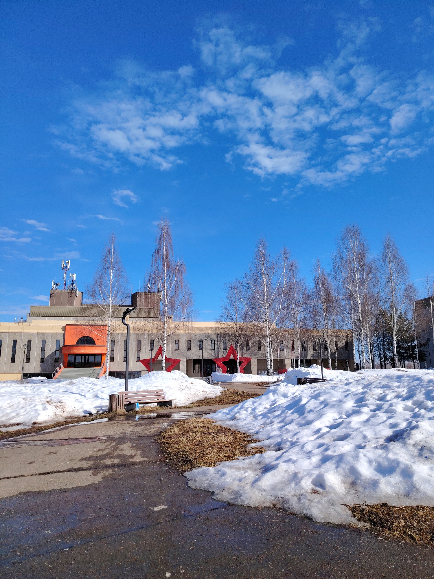 The image presents an outdoor scene in Марковский, Russia, under a clear blue sky dotted with scattered white clouds, indicative of a sunny late winter or early spring day. A multi-story building, constructed with gray concrete panels and horizontal beige sections, occupies the midground. The building features numerous vertical rectangular windows. On the left section of its roof, a cluster of telecommunication antennas and equipment is visible. A prominent orange entrance with a semi-circular arch is centrally positioned, accessible via concrete steps. Two large, red, five-pointed stars are affixed to the building's facade; one is located to the right of the orange entrance, and another is further right, partially obscured by trees.
The foreground consists of a dark, wet asphalt or concrete path that curves from the lower left towards the center, displaying puddles and reflective surfaces. Large, irregular mounds of melting snow cover the ground, interspersed with patches of exposed brown, dry grass. Numerous bare birch trees with white bark and slender branches are distributed across the midground in front of the building, with additional coniferous trees visible in the far right background. A single wooden park bench, featuring three dark horizontal slats, is situated to the right of the path, partially encompassed by snow. No individuals are present in the depicted environment.