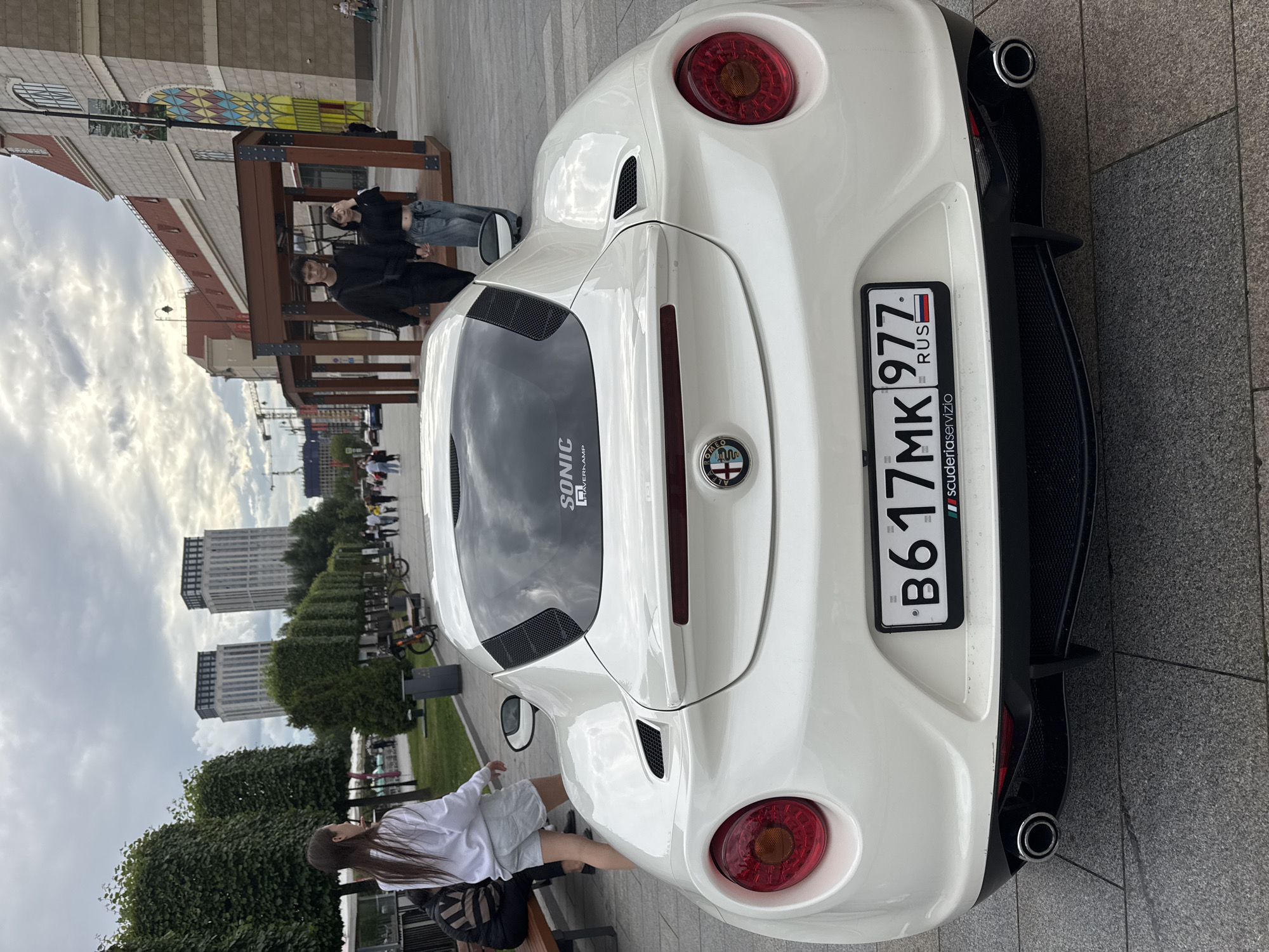 A white Alfa Romeo 4C Spider is parked on a gray paved ground in front of a building with a red roof. The car's license plate reads "B617MK 977 RUS". In the background, two young people are walking, holding hands. Behind them, a wooden structure with a canopy is visible, and further back, two tall buildings with dark roofs stand against a cloudy sky. The scene appears to be in Moscow, Russia, with the architecture and style of the buildings suggesting an urban environment. There are trees and streetlights present in the middle ground, and a woman in a white top and light shorts is walking away from the camera on the left.