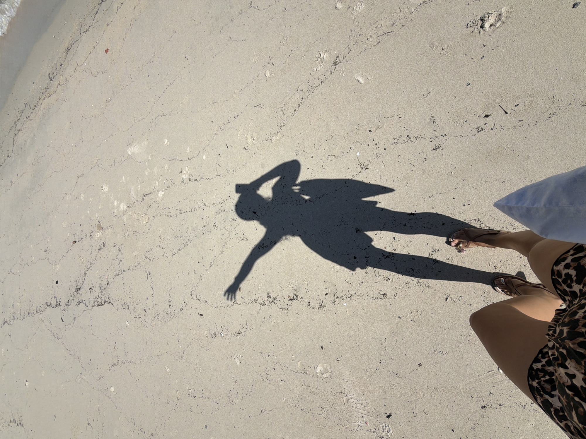 A shadow of a person is cast on a sandy beach. The shadow shows the person holding a mobile phone and a bag. The person is wearing a dress and sandals. The beach is wet in some areas, indicating it is close to the water. The sand has some debris and footprints on it. The scene is likely in Ko Samui District, Thailand.