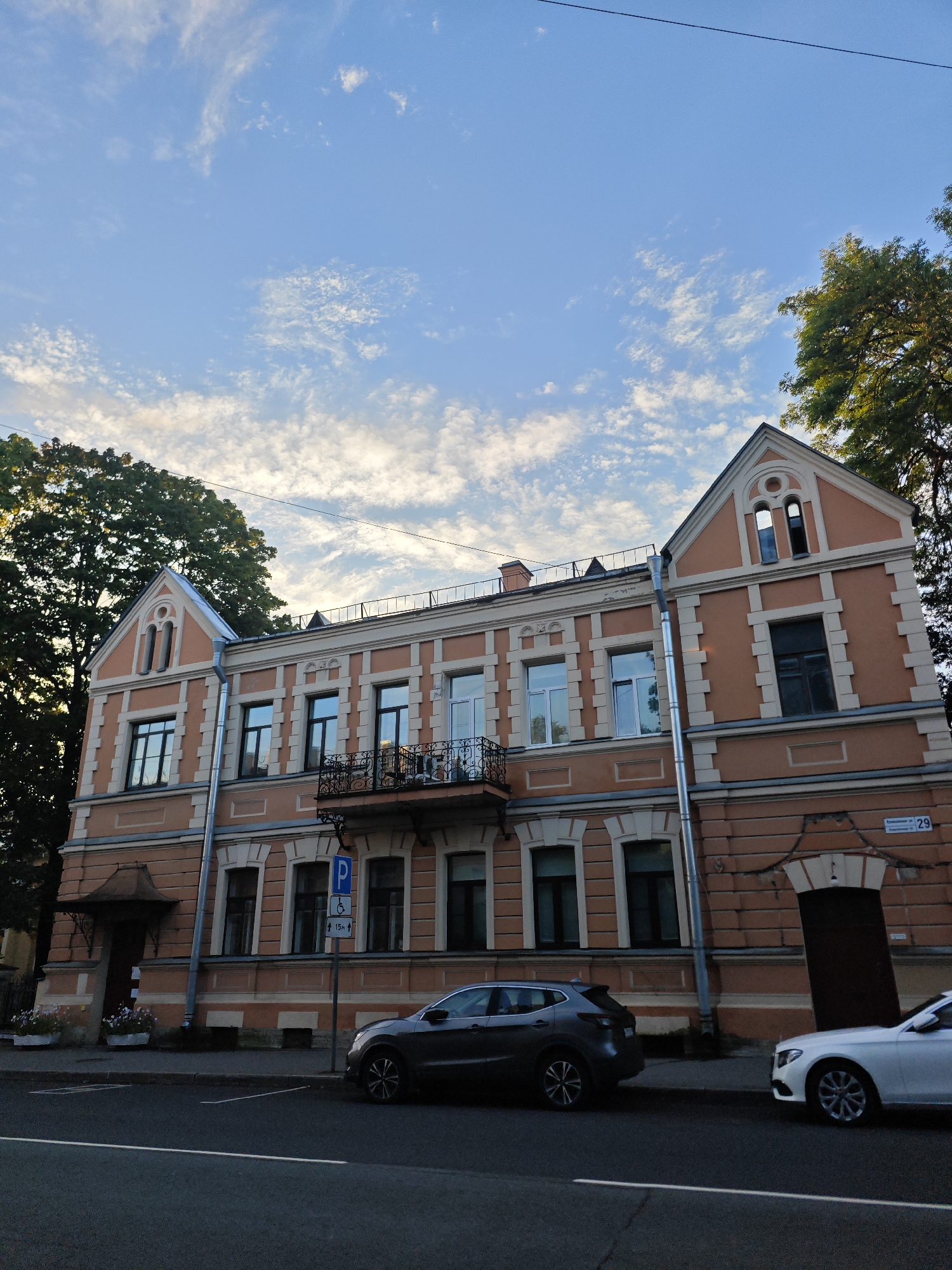 The image captures a street scene in Pushkin, Russia, featuring a three-story salmon-colored building with ornate architectural details, including decorative window surrounds, a balcony, and a gabled roofline. The building is flanked by mature trees. In front of the building, a dark grey SUV is parked on the street, and a white sedan is parked further down the street. A blue parking sign with an accessibility symbol is visible near the building. The sky above is a clear blue with scattered wispy clouds. A portion of a street with a white lane marker is in the foreground.