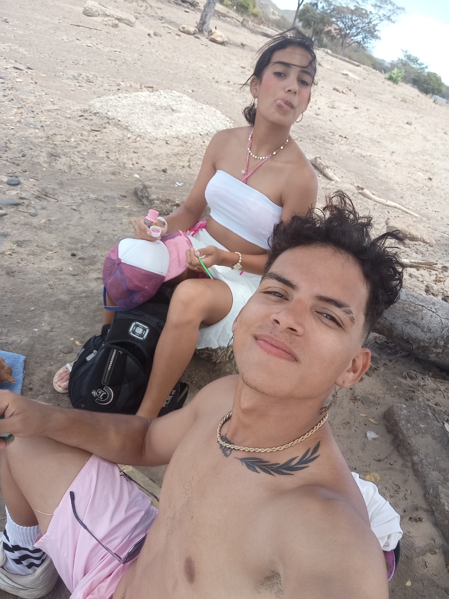 Two individuals are posing for a selfie at a beach in Altagracia, Venezuela. The foreground focuses on a shirtless male wearing pink shorts, white socks with black stripes, and sneakers. He has a gold chain necklace and a laurel branch tattoo on his left clavicle. The background features a seated female wearing a white tube top, white skirt, and a pink necklace. She is sticking her tongue out and holding a pink bottle. She is seated on top of a black backpack. A pink and white baseball cap is also visible. The setting appears to be a beach with light-colored sand and sparse vegetation.