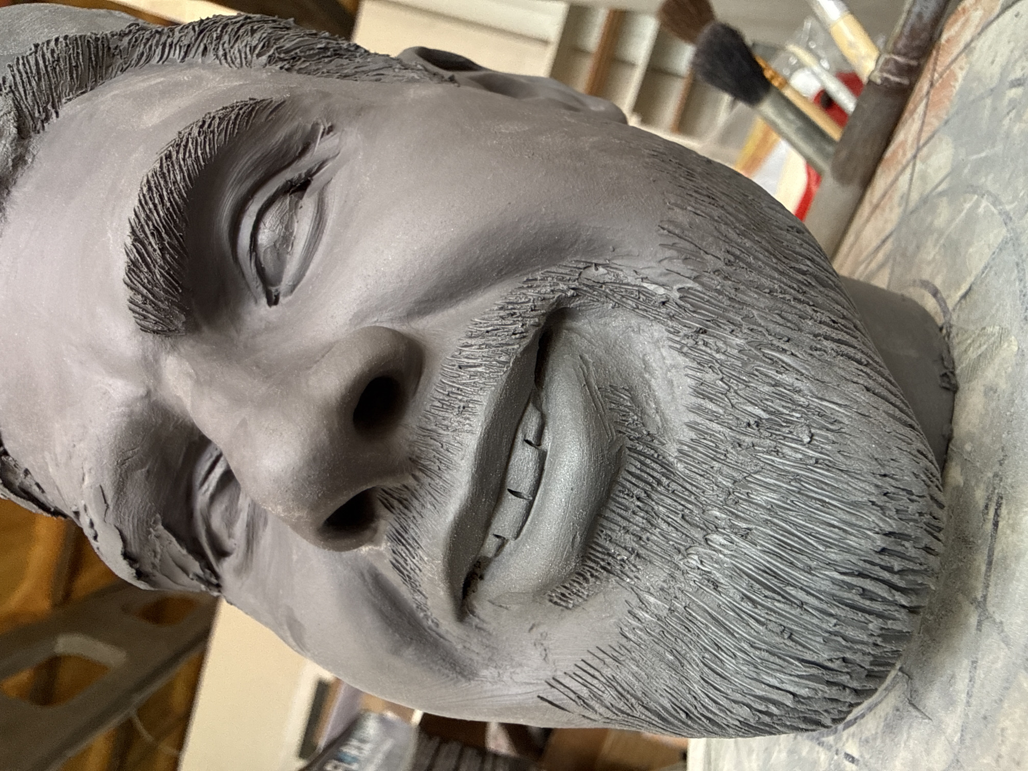 A gray clay sculpture of a man's head and shoulders is visible. The sculpture is on a surface, near some painting brushes. The man's eyes are closed and he appears to be smiling. There is no indication of a location in the city of Chaikovsky, Russia.