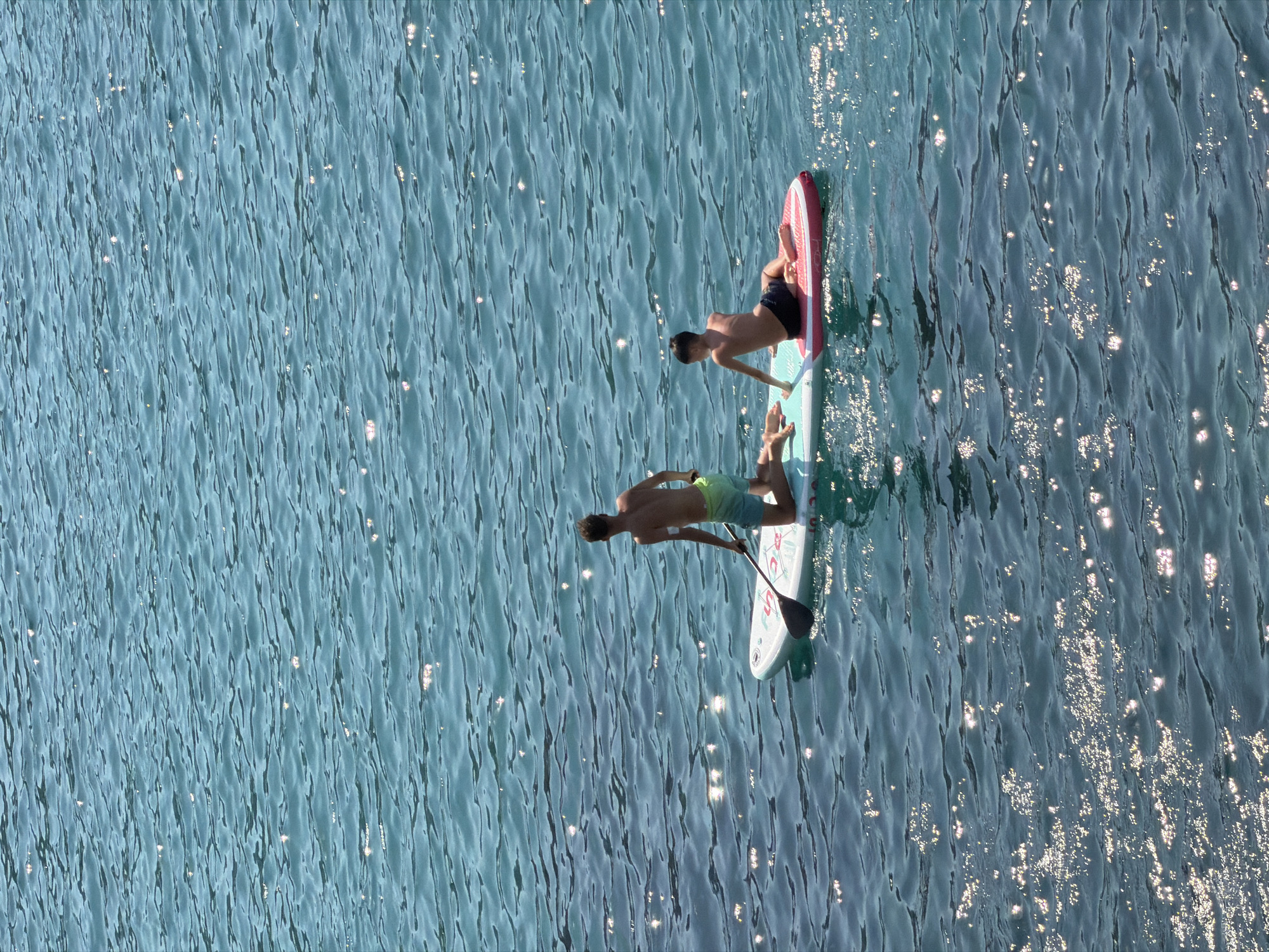 Two young males are paddleboarding on the clear, blue waters of Bečići, Montenegro. The person in the foreground is kneeling on the paddleboard, holding a paddle and looking forward. The second person is lying down on the paddleboard. The sun is shining, creating a shimmering effect on the water.