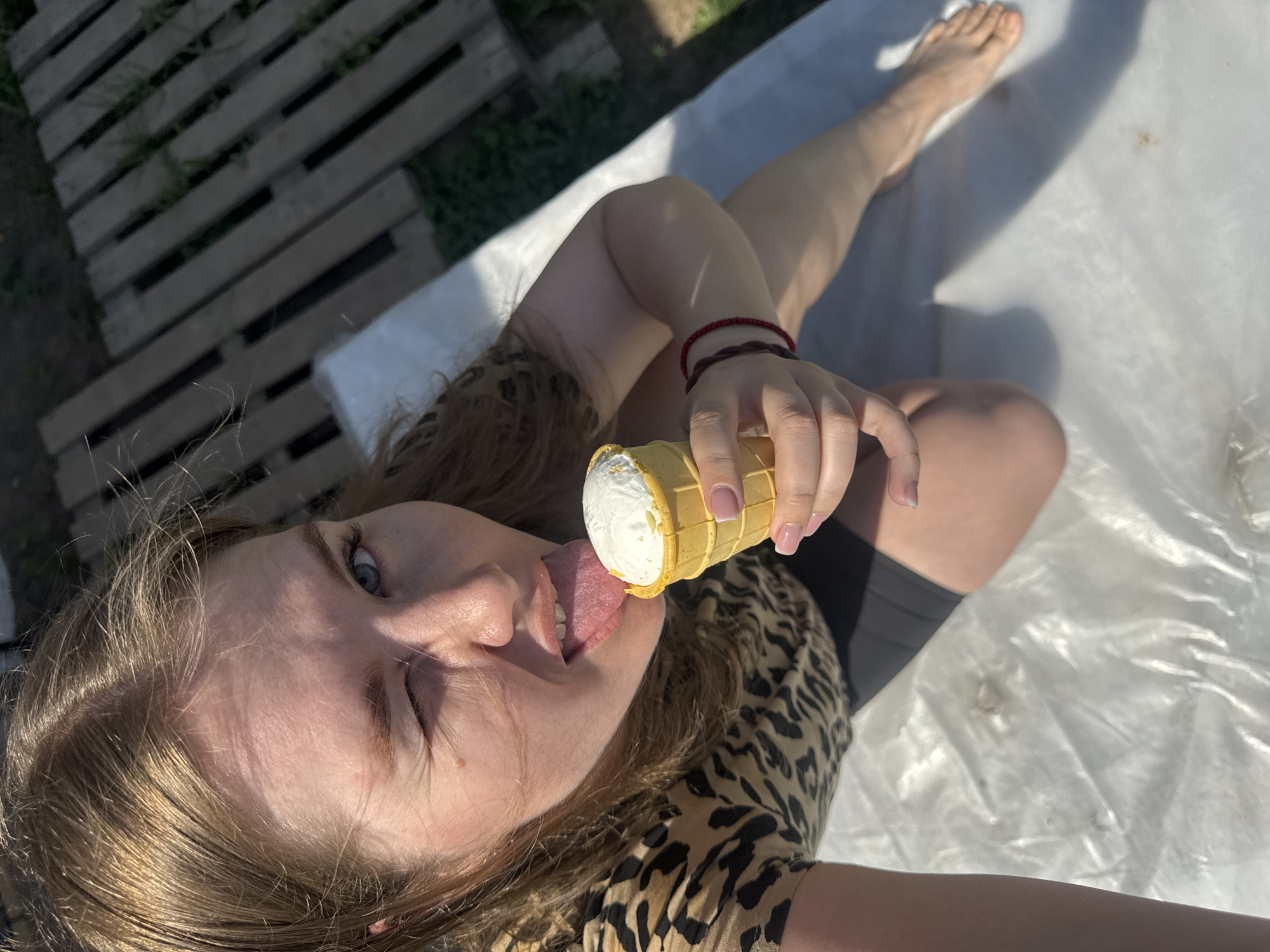 A blonde-haired Caucasian woman with blue eyes is lying on a white, possibly plastic, sheet outside in Ферма, Russia. She is wearing a leopard-print shirt and black shorts. Her right leg is extended towards the upper right corner of the frame, revealing her bare foot. Her left arm is extended towards the camera, holding an ice cream cone with white ice cream. The woman is licking the ice cream with her tongue extended and her right eye closed in a squint. On her right wrist are two bracelets, one red and one dark brown. To the left, a section of a wooden pallet is visible with grass growing between the slats. The overall lighting suggests it is daytime.
