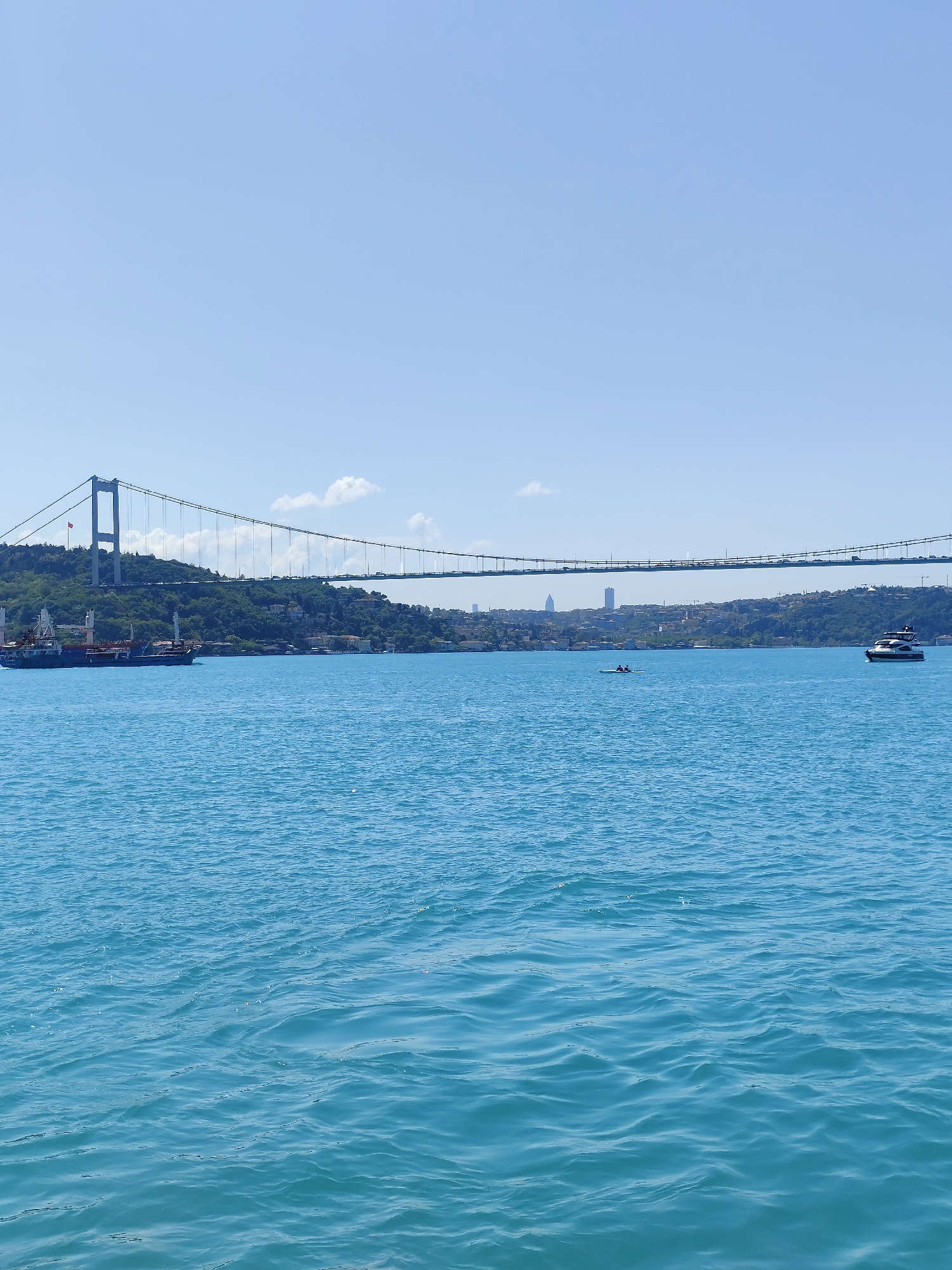 The image depicts a view of the Bosphorus Bridge in Sarıyer, Turkey, under a clear blue sky. The bridge spans the waterway, connecting two landmasses covered in green vegetation and scattered buildings. In the water, a cargo ship is situated on the left, a small rowboat with two occupants in the center, and a motor yacht on the right. The surface of the water is rippled with gentle waves.