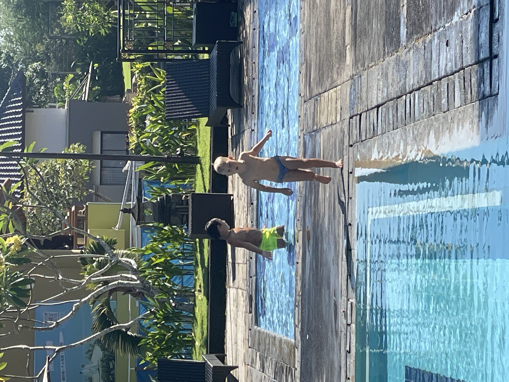 Two young children are present in an outdoor swimming pool area. On the left, a child wearing neon green swim trunks stands in a shallow section of the pool, facing away from the camera and towards the right. On the right, a child wearing blue swim briefs is in motion, running along the wooden decking adjacent to a deeper section of the pool. The background features lush green foliage, lounge chairs, and part of a building with a dark roof and beige exterior walls, suggesting a resort or hotel environment in Kuta Selatan, Indonesia. The scene is brightly lit by sunlight, casting shadows on the wooden decking and the water's surface.