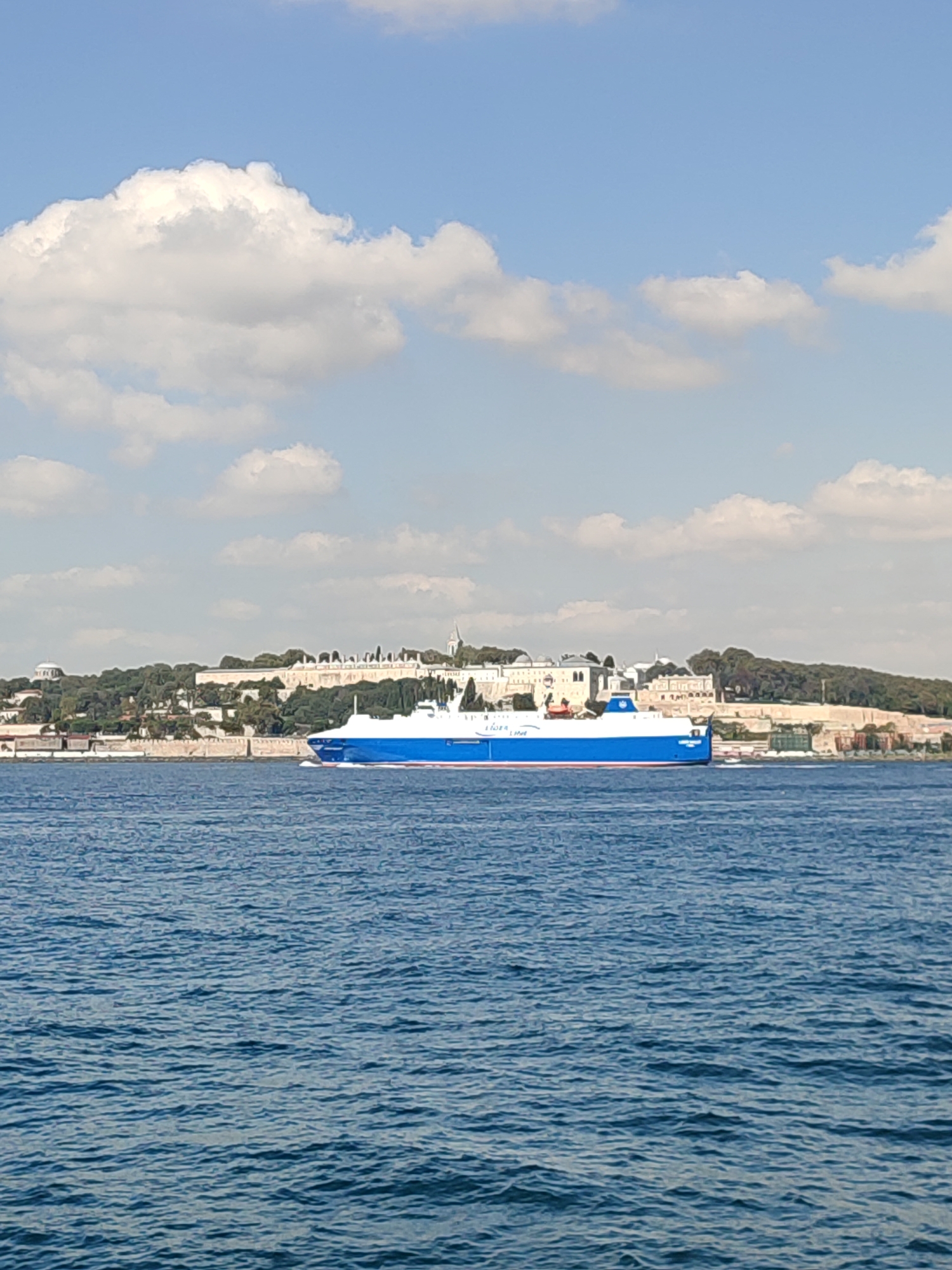 A wide shot captures a large, blue and white ferry sailing across a body of water, likely the Bosphorus Strait, with a historical cityscape on the horizon. The ferry, prominently in the foreground and midground, is a long vessel with a white superstructure and a bright blue hull. The cityscape in the background features a sprawling complex of light-colored buildings situated on a green, tree-covered hill. The architecture suggests historical significance, with multiple domes and structures visible. The sky above is a clear blue, dotted with fluffy white clouds. The water is a deep blue, with gentle ripples reflecting the sunlight. The scene is tranquil and depicts a moment of transit against a backdrop of history and natural beauty.