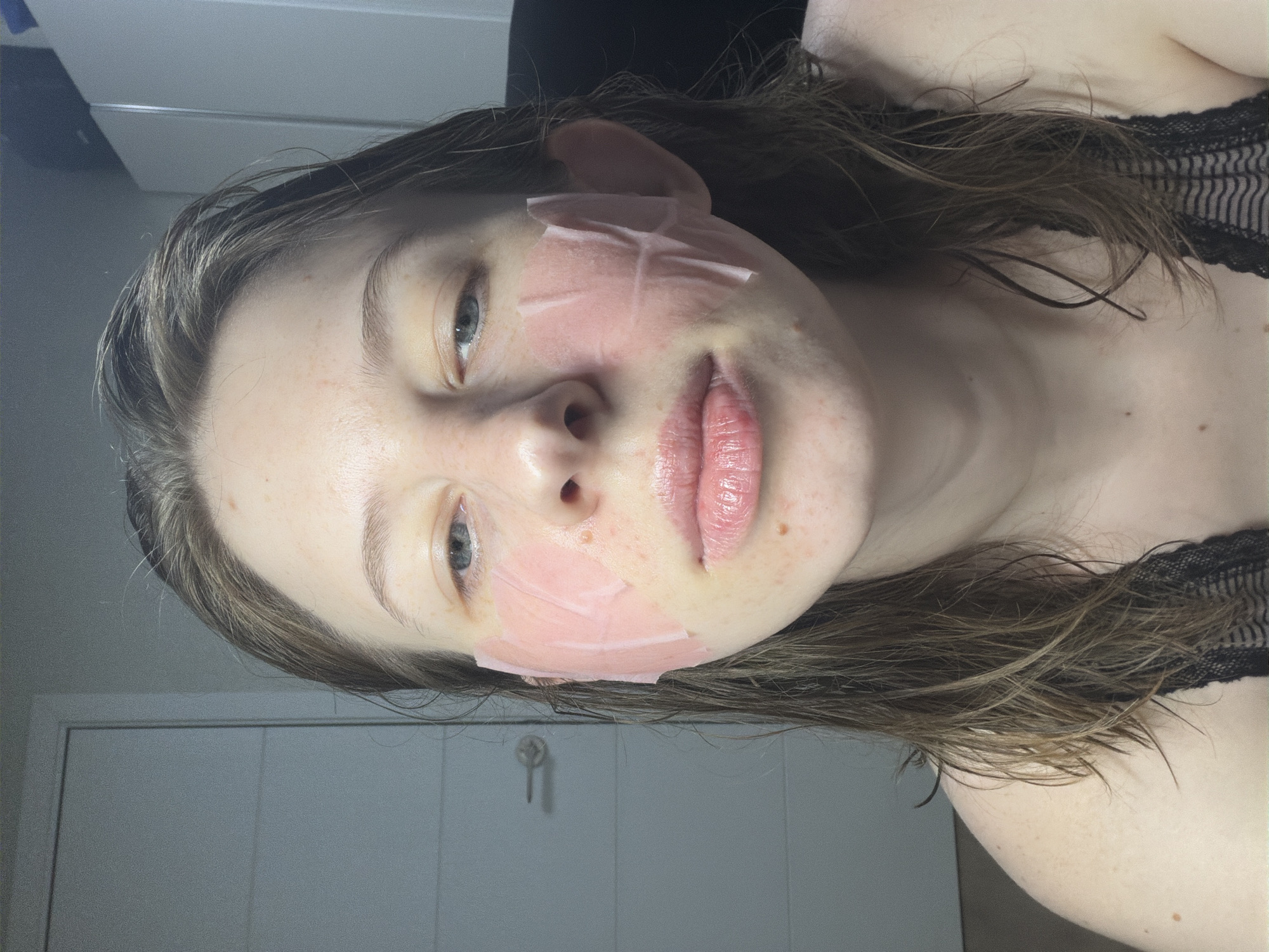 A young woman with light brown, wet hair and blue eyes is in a close-up portrait, facing forward. She is wearing a black lace top. Pink under-eye patches are applied to her cheeks. She has light skin with freckles and some redness. The background appears to be an indoor setting with a white door on the left and white cabinets on the right, suggesting a residential environment in Dongducheon-si, South Korea.