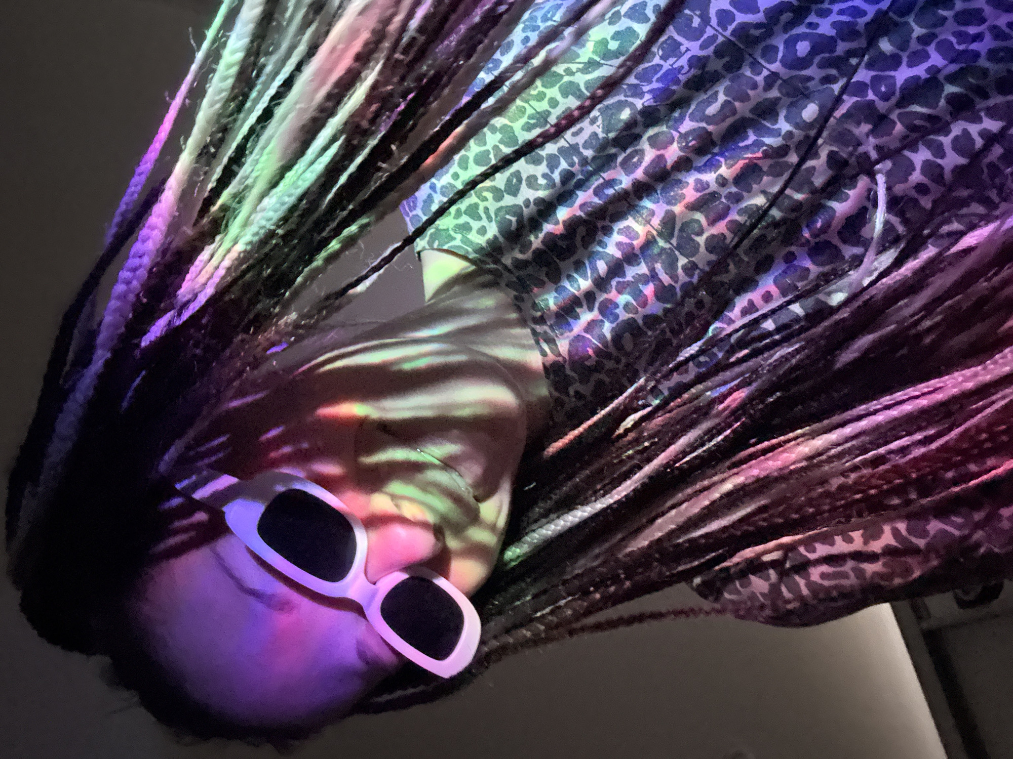 The image shows a person with braided hair, wearing white sunglasses, and an animal print shirt. The person is likely indoors. The lighting is colored with pink, green, and blue hues. The person is looking down. The image is rotated counterclockwise. There is no indication of the location being Rusanovo, Russia.