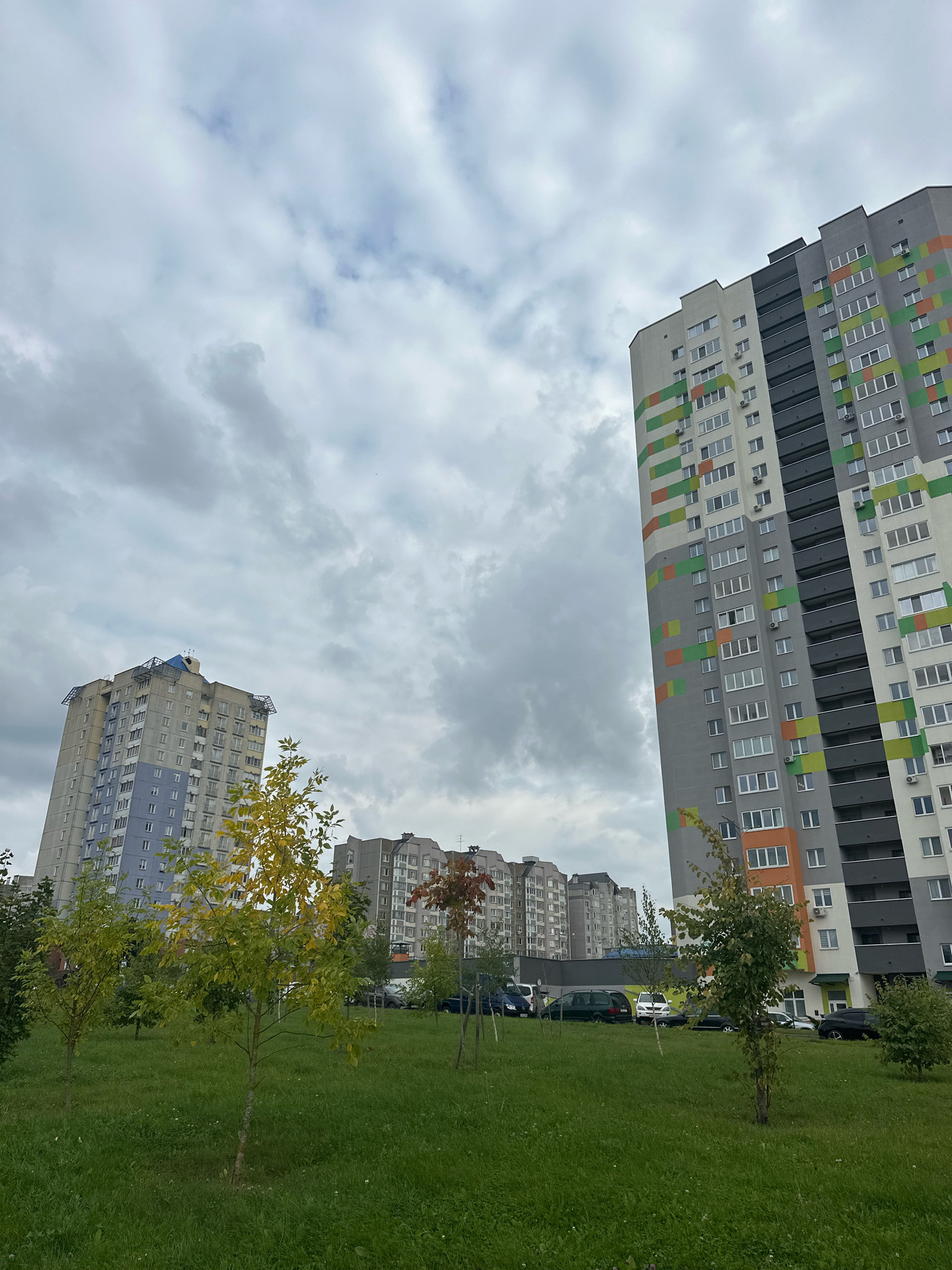 A low-angle outdoor shot shows a park area in front of several modern apartment buildings under a cloudy sky. In the foreground, a grassy area is dotted with young trees, some with yellowing leaves, and in the middle ground, several cars are parked. Behind the trees and cars, three apartment buildings are visible. The closest building, on the right, is tall and grey with white, green, and orange rectangular patterns on its facade. To its left and further back is another tall apartment building, colored light blue and grey. The furthest building, in the center-left, is also a multi-story apartment building, grey with patterned balconies. The sky is overcast with dense, grey clouds. The scene appears to be in an urban residential area, likely on an autumn day. This scene is located in the Minsk World complex in Minsk, Belarus.