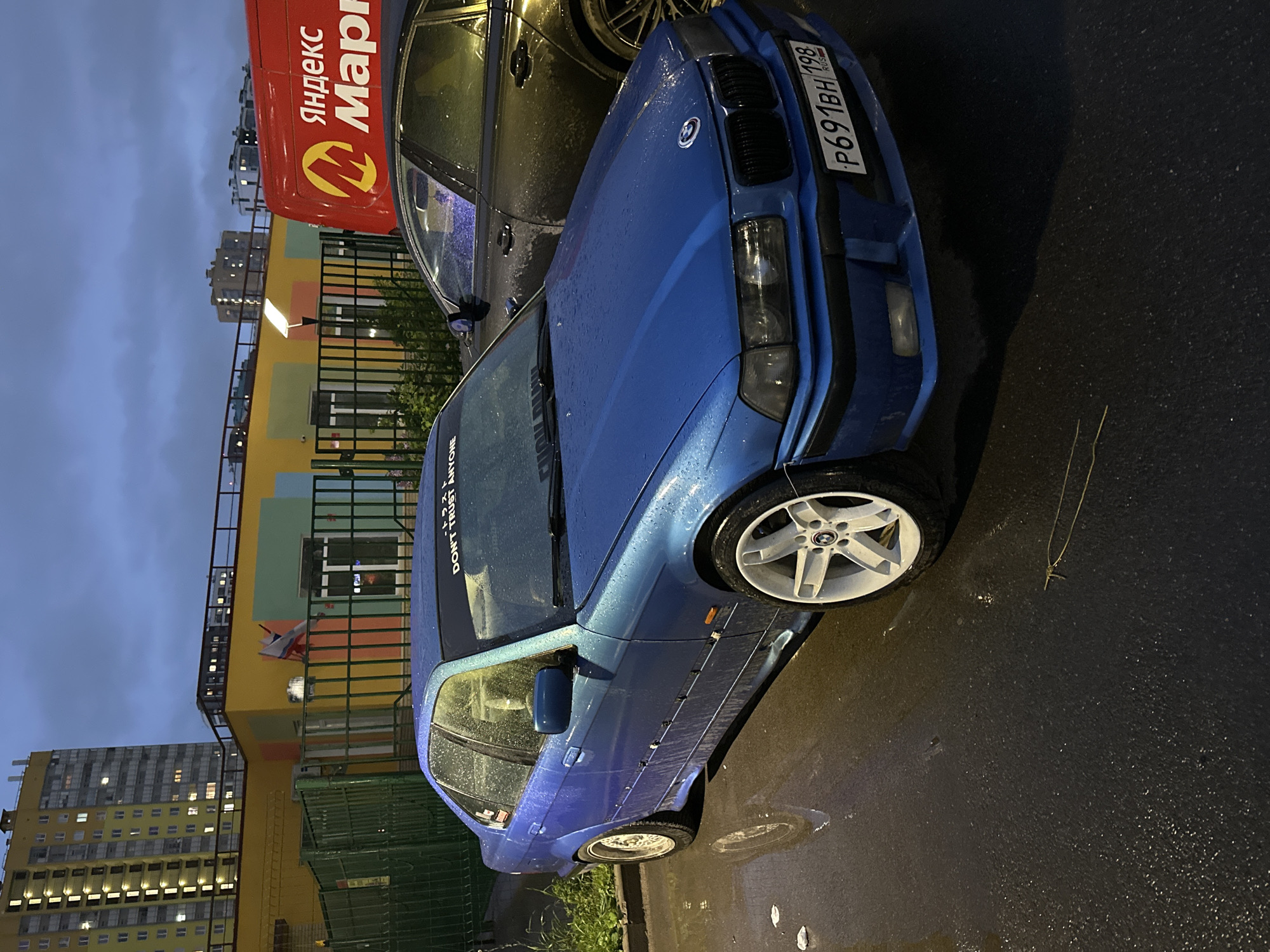 The image shows a blue BMW E36 parked on a wet street. Its license plate is P691BH 198, indicating it is registered in Russia. A black car is parked next to it, partially visible. The street appears to be in a residential area, with a high-rise apartment building visible in the background. A sign for "Yandex Market" is visible on a building. Based on these visual elements, the location of the image is identified as Murino, Russia.