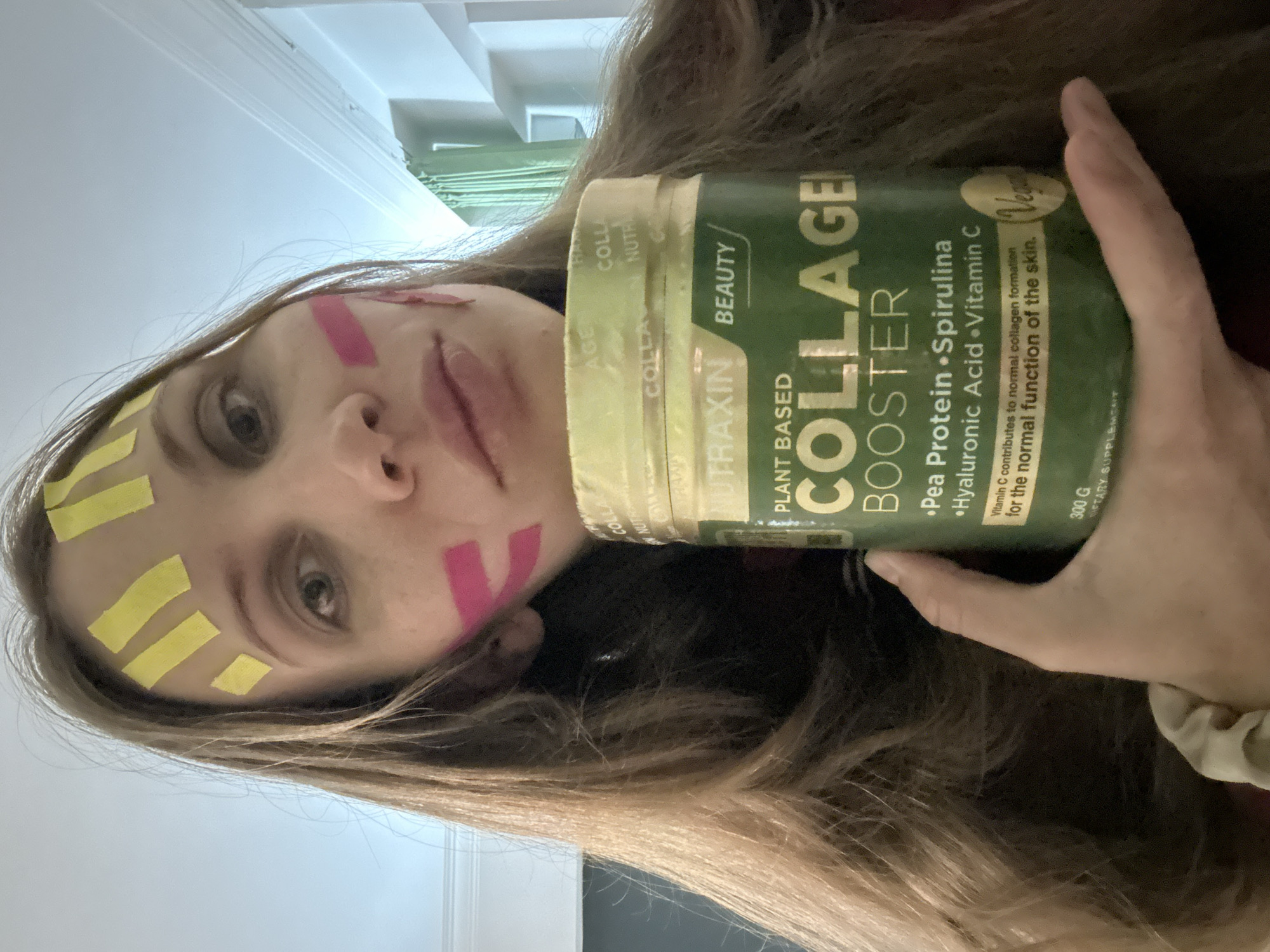 A woman is holding a container of "Collagen Booster" by Nutraxin Beauty, which is a plant-based product containing pea protein, spirulina, hyaluronic acid, and Vitamin C. She has yellow and pink tape on her forehead. The location is unknown, but the city of Fethiye, Turkey has not been established.