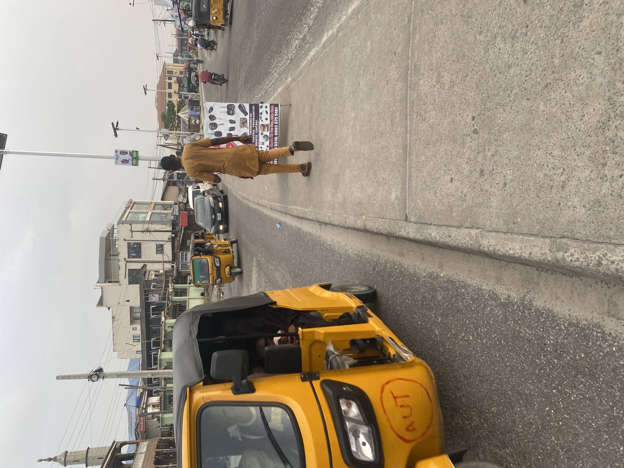 The image depicts a street scene in Ado, Nigeria. A man in a tan-colored outfit is walking past a sign advertising key programming services. The sign provides a phone number and identifies the location as Oke-Ira Nla Bus Stop, Addo Road. A yellow auto rickshaw, or "keke", is in the foreground, with the letters "AUT" written in red inside a circle on the vehicle's side. In the background, more kekes and cars are visible. Buildings and utility poles line the street.