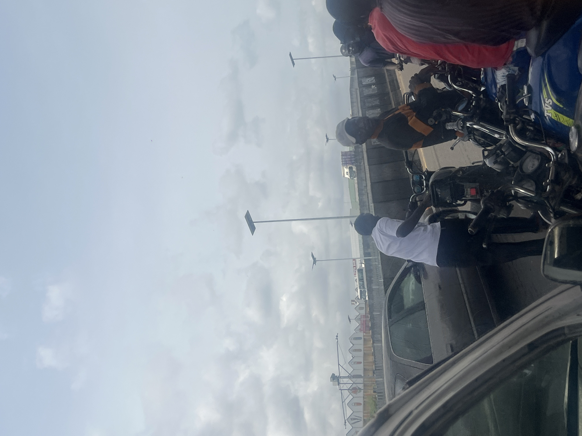 The image depicts a traffic scene in Agani, Nigeria, likely on a bridge or elevated road. The sky is overcast. Motorcycles are densely packed on one side, with riders wearing helmets. Various colors of shirts are visible on the riders. To the left, a row of white structures with arched roofs is seen. Cars are also present, with the photo appearing to be taken from inside a vehicle. Streetlights line the road. The scene suggests traffic congestion.