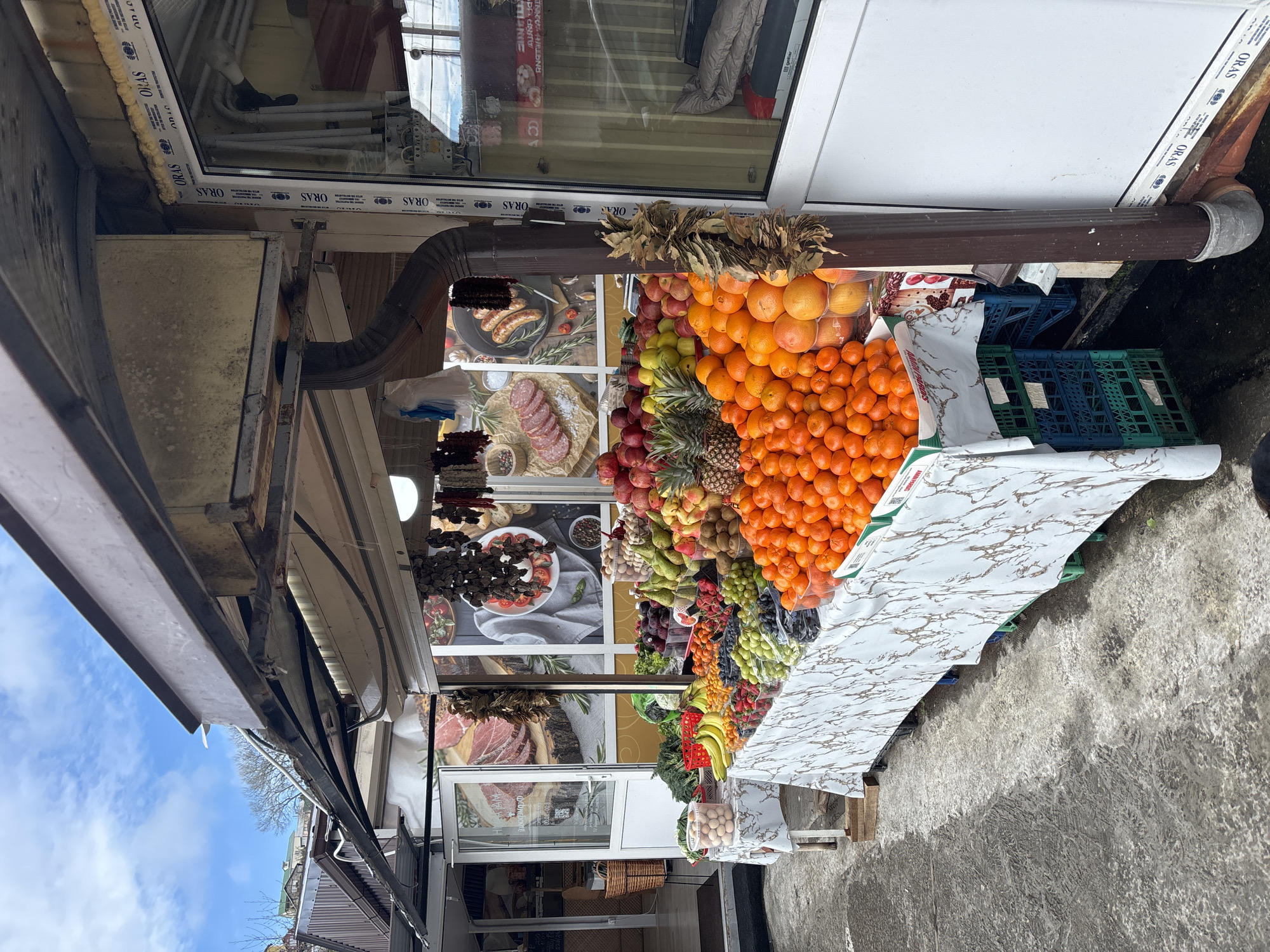 A fruit stand is depicted in a photograph, most likely taken in Sochi, Russia, based on the text in the image. The stand displays a variety of fruits, including oranges, mandarins, pineapples, grapes, apples, pears, bananas, and possibly other produce. The backdrop behind the fruit features printed images of various foods. The stand itself has a white marble-patterned countertop and appears to be located adjacent to a building with a window, and an awning structure overhead. The ground surface in front of the stand is concrete.