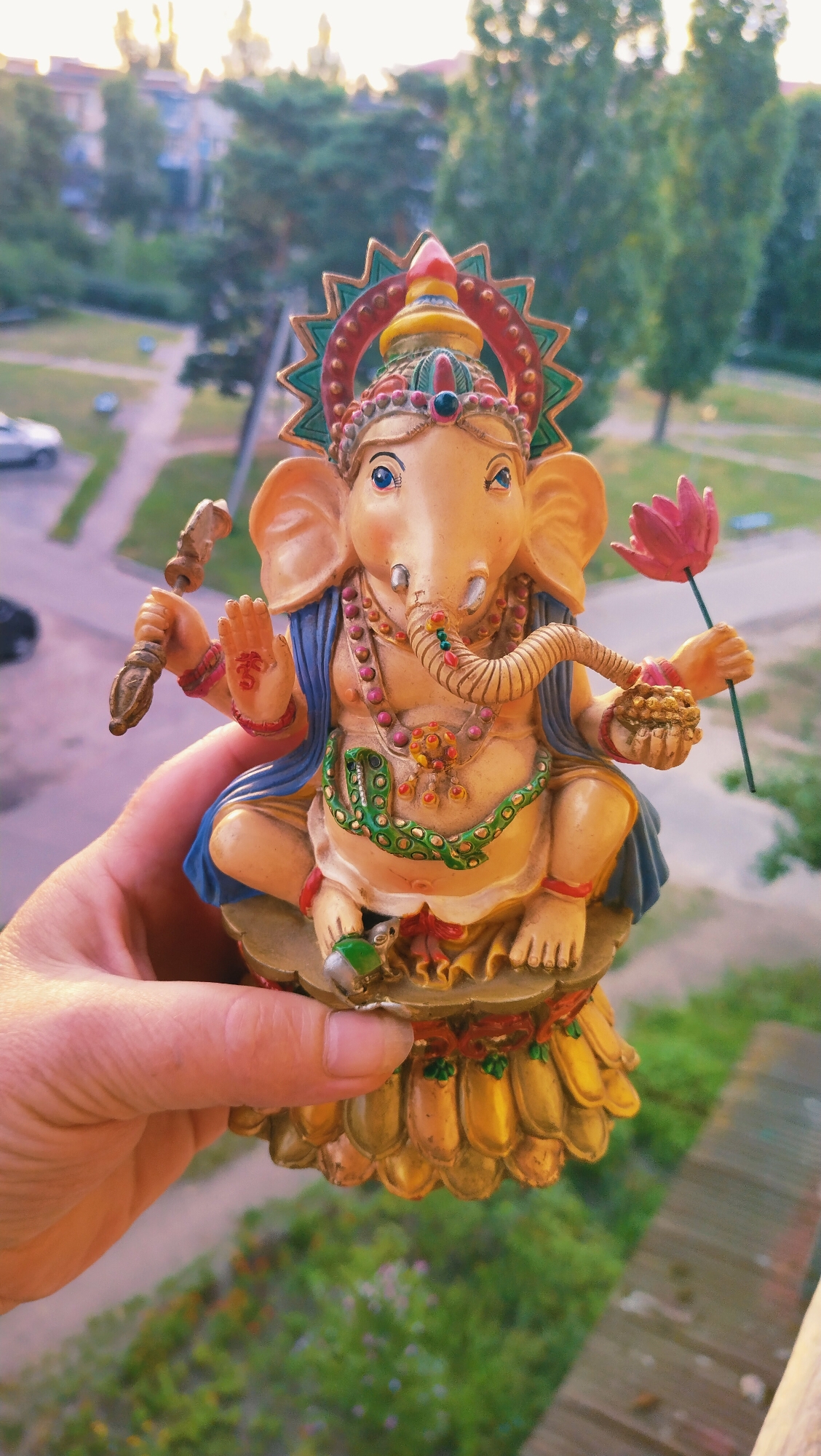 A hand holds a statue of Ganesha, the Hindu deity, outdoors. The statue depicts Ganesha seated, with an elephant head, blue eyes, and multiple arms. He wears ornate jewelry, including necklaces and a green serpentine design around his torso. One hand holds a pink lotus flower, while another holds a broken pestle. The statue is colorful, with gold, red, blue, and green accents.
The background suggests an urban environment with buildings and trees, consistent with the city of Horishni Plavni, Ukraine. The setting appears to be a balcony or elevated area, as indicated by the railing in the lower right corner. A blurred out car is visible on a road in the mid-ground. The lighting suggests it is daytime, likely late afternoon or early morning due to the warm tones of the sunlight. The focus is on the statue and the hand holding it.