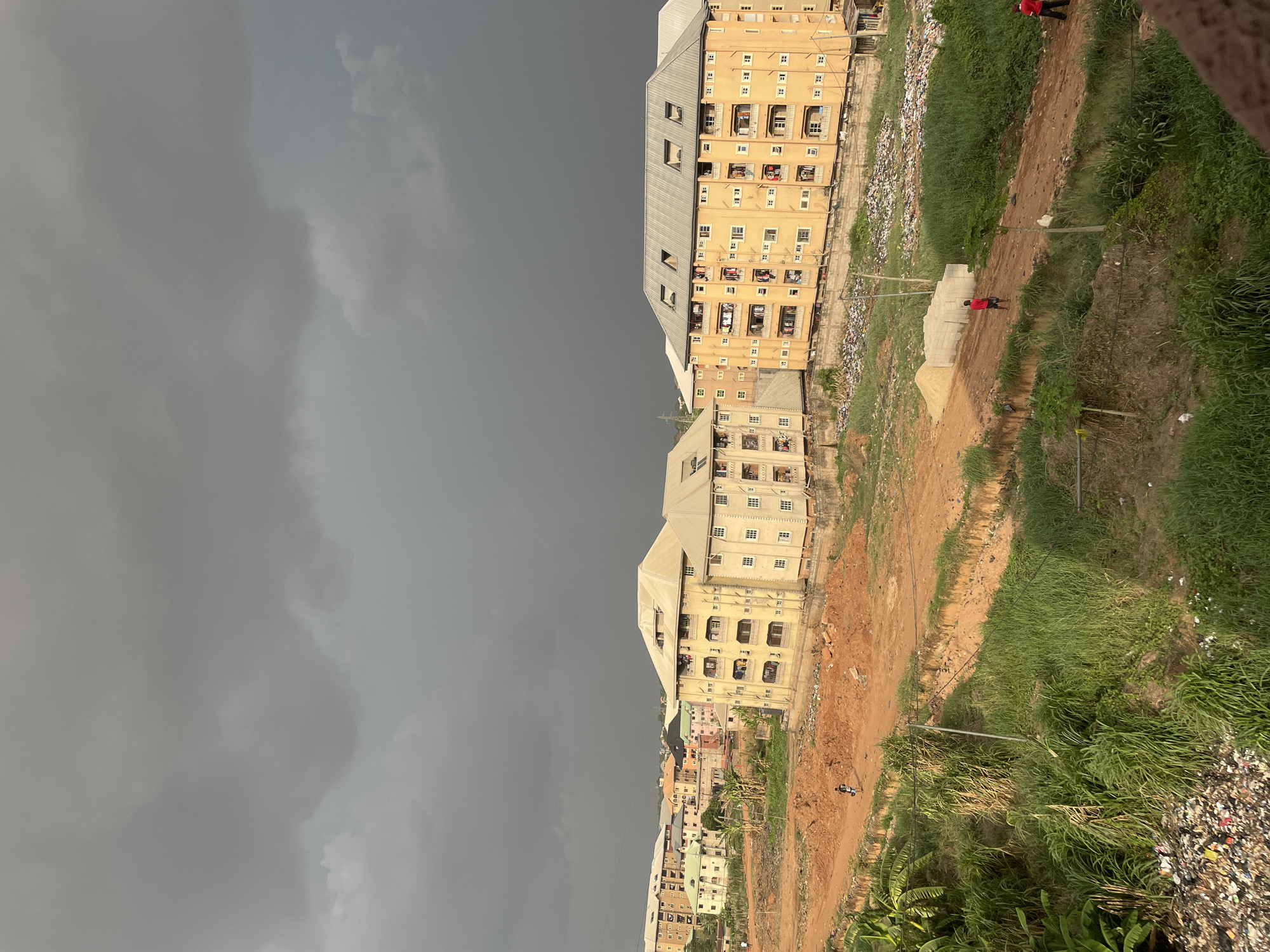 The image shows an urban scene in Onitsha, Nigeria, under a heavy, overcast sky. Several multi-story residential buildings are visible, characterized by yellow facades, metal roofs, and numerous windows and balconies. An unpaved road runs adjacent to the buildings, bordered by dense vegetation on one side and accumulated garbage on the other. Two people wearing red shirts are visible in the scene.