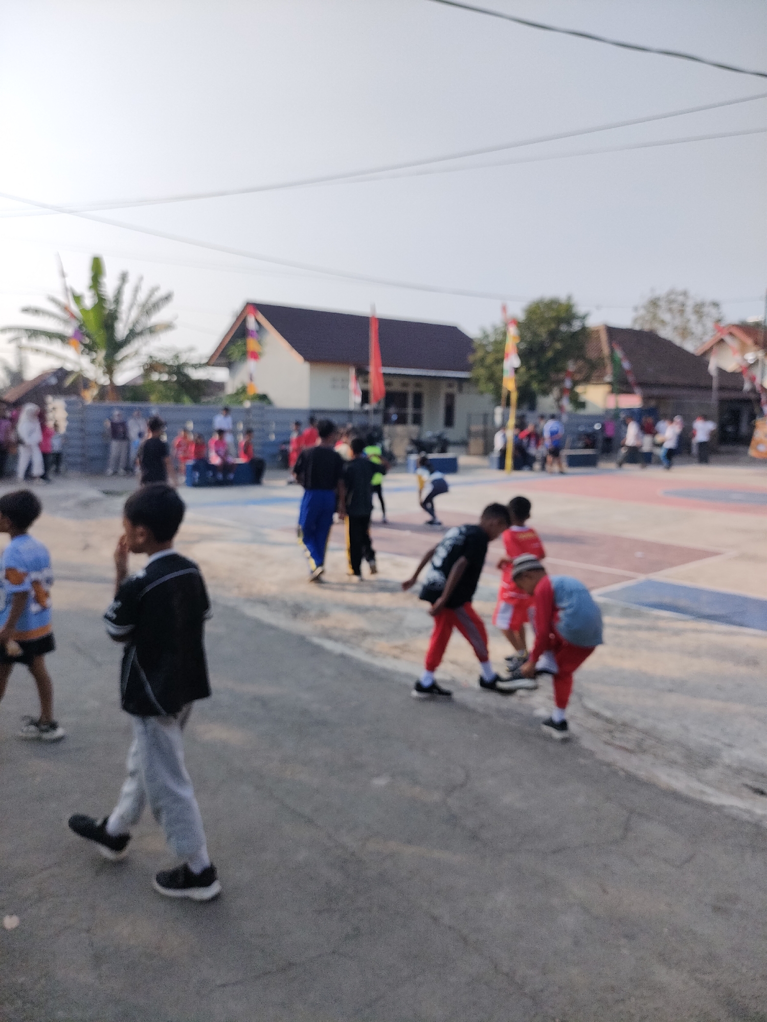 The image depicts an outdoor scene with several children and adults. The setting appears to be a schoolyard or community gathering area, with buildings and a paved court visible in the background. Some of the children are engaged in physical activity, while others are walking or observing. The overall atmosphere is casual and appears to be daytime. The location is specified as Caracas, Indonesia.