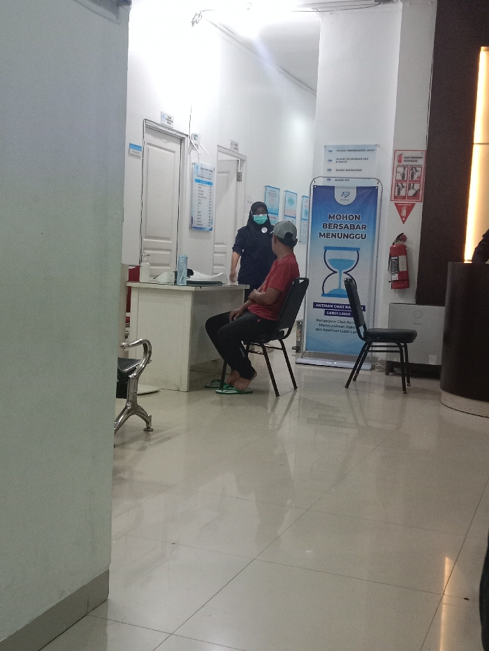 The image depicts an indoor scene within a medical facility or clinic reception area in Gladaksari, Indonesia. A masked female staff member, wearing a dark blue uniform and a black head covering, stands behind a white desk. She faces a male patient seated on a dark chair in front of the desk. The male wears a red t-shirt, dark trousers, and a grey baseball cap worn backward; his bare feet rest on light green flip-flops placed on the floor. His arms are crossed.
On the white desk, a hand sanitizer dispenser, a white container, and papers are visible. The background features light-colored walls with multiple white doors and various blue and white informational signs, including one indicating "RUANG KAI" (KAI Room). A prominent freestanding blue banner on the right displays the Indonesian text "MOHON BERSABAR MENUNGGU" (Please Be Patient While Waiting) with an hourglass graphic, and below it, "ANTRIAN OBAT RAHI" (Drug Queue) with additional text on wait times. To the right of the banner, a red fire extinguisher is mounted on the wall below a pictogram-based fire action sign. The floor is covered with reflective light-colored tiles, showing reflections from overhead lighting. A dark, cylindrical object is partially visible on the far right, and the base of a chrome chair is seen on the far left foreground.