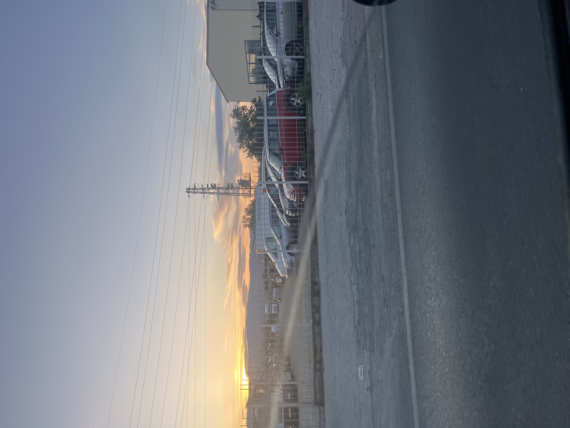 The image shows a street in Blagoevgrad, Bulgaria at sunset. A row of parked cars sits behind a metal fence, next to a building with a light-colored facade. Overhead electrical lines stretch across the sky. The road is in the foreground, with the light of the setting sun visible in the background.