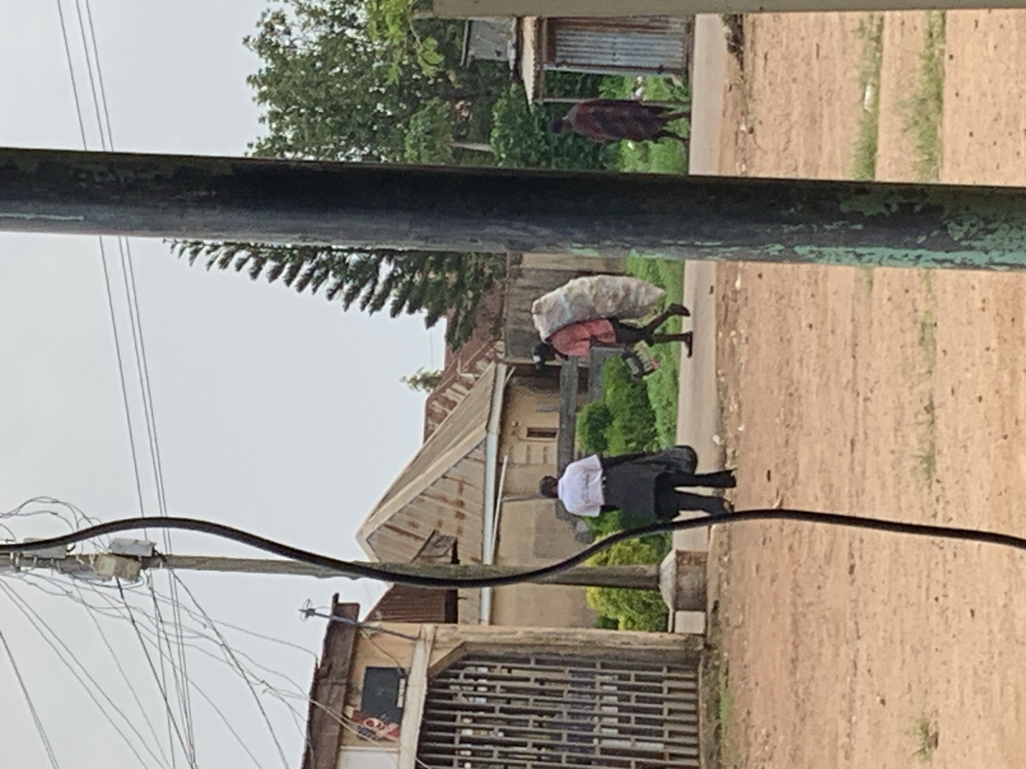 The image depicts an outdoor scene in Jos, Nigeria. In the foreground, a dark pole with peeling green paint is positioned on the right side. Behind it, a dirt road runs horizontally across the frame, with a person in a white shirt and dark pants carrying a black bag walking away from the camera on the left side. Further back, a man wearing a pink shirt and dark shorts carries a large, light-colored bundle of material on his shoulder, walking towards the right. To the far right, another person is seen walking away, wearing a dark red and brown patterned garment. The background features several buildings with corrugated metal roofs, a large evergreen tree, and dense green foliage. Overhead, numerous electrical wires are strung between poles. The sky is overcast.