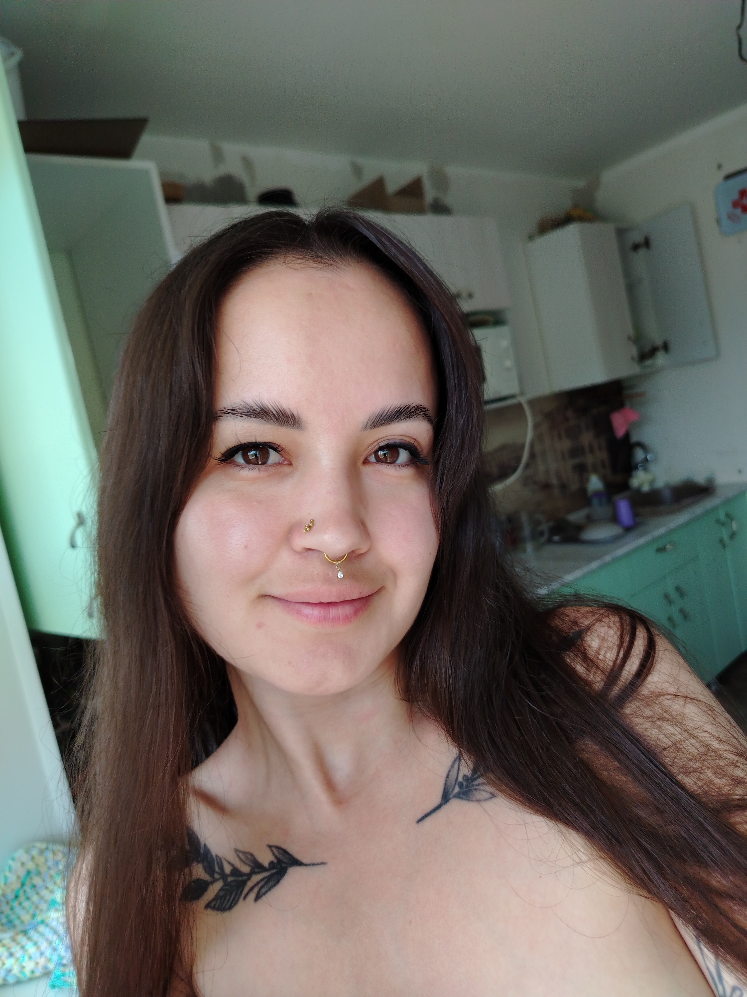 A close-up shot of a fair-skinned woman with long brown hair, with two visible tattoos near her collarbones: one of a branch with leaves and another of a simple floral design. She has dark eyebrows, brown eyes, and a light complexion. She is wearing a gold nose ring and a stud in her nostril. The background shows a kitchen setting with white cabinets and green countertops. The location information is not visually discernible and, therefore, is not part of the description.
