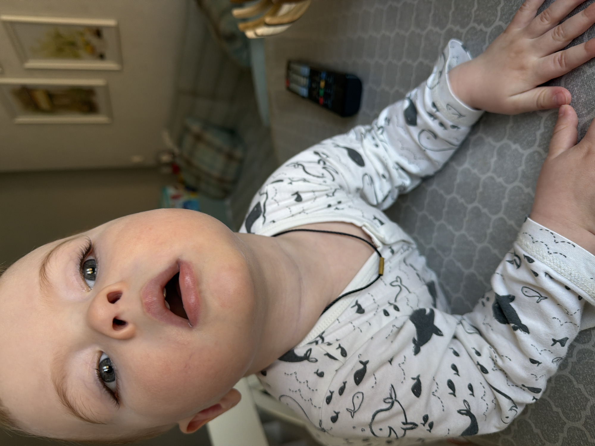 A close-up, high-angle shot depicts a baby lying on a grey surface. The baby is wearing a white onesie patterned with grey marine animals. The baby's mouth is open. The background includes a black remote control, a patterned cushion, and framed pictures on a wall. The location is not identifiable as Votkinsk, Russia from the visual information.