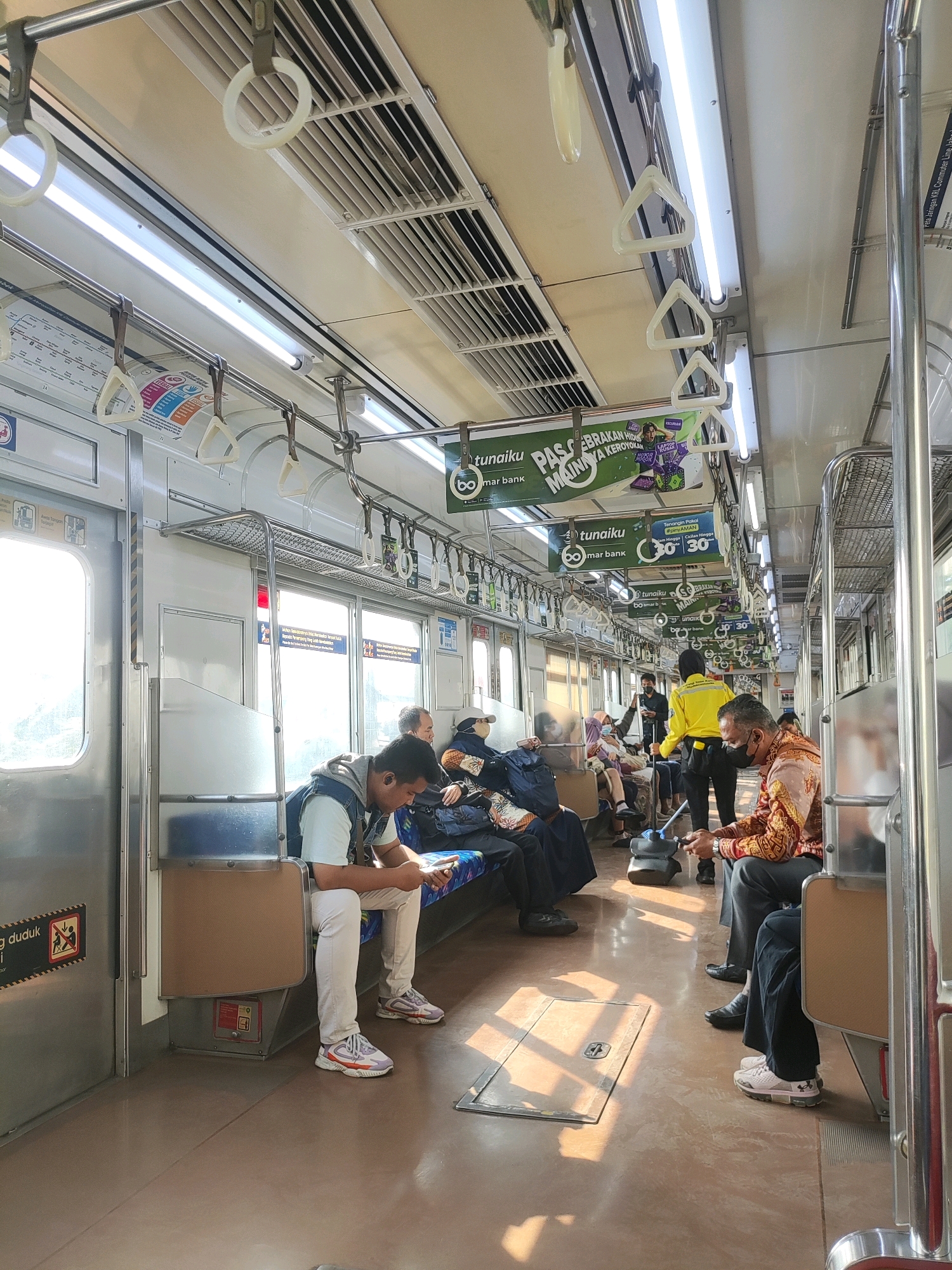 This is a photograph taken inside a commuter train, likely in Bekasi, Indonesia, given the context of the question. The train is populated with several passengers, both seated and standing, though mostly seated. The interior is characterized by cream-colored walls and ceiling, with fluorescent lighting illuminating the space. Silver grab bars and white plastic handles for passengers to hold onto are visible throughout the carriage.
In the foreground, a young man wearing a denim vest over a white t-shirt and white pants is seated, engrossed in his mobile phone. To his right, further down the aisle, several other passengers are seated, including a woman wearing a hijab and a man in a patterned shirt. A train conductor or attendant in a bright yellow jacket and black pants is standing and attending to passengers, holding a cleaning tool. Another man, wearing a mask and a patterned shirt, is seated in the right foreground, also looking down. The floor is a neutral brown color, with sunlight casting distinct rectangular patterns of light and shadow. Several advertisements are mounted on the ceiling and walls, with visible text in Indonesian. The overall impression is of a typical commuter train journey, with passengers engaged in their own activities.