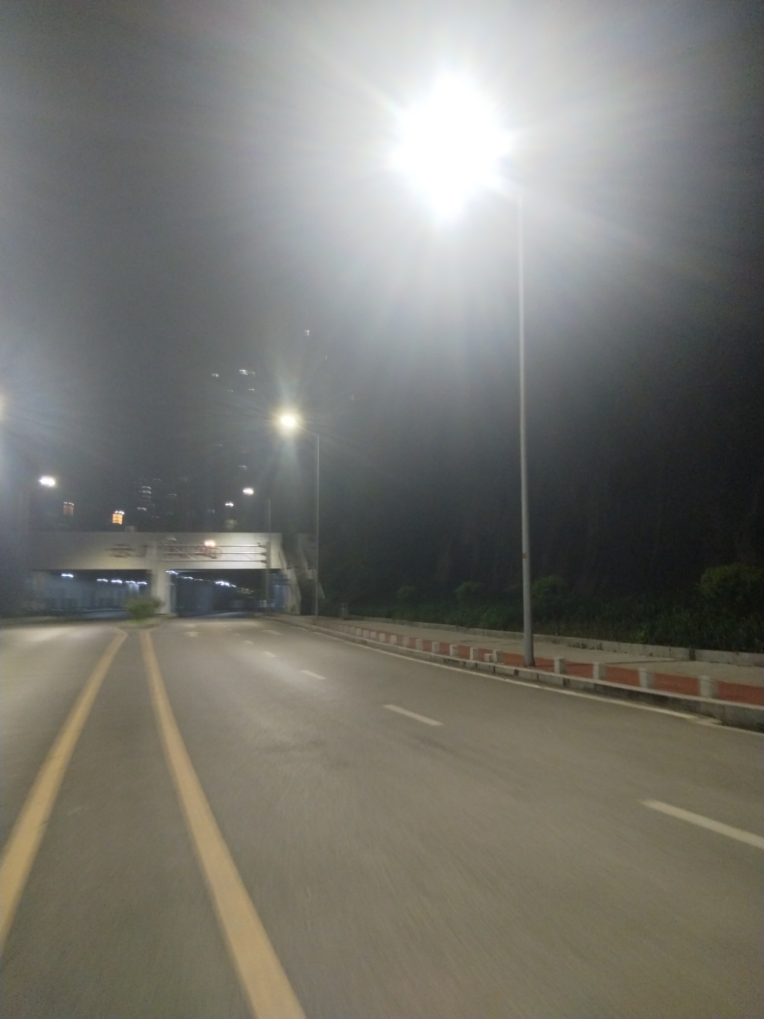 The image captures a night scene on a road in Jiulongpo District, China. The street is illuminated by bright overhead streetlights that create a strong glare. A tunnel structure spans the road, with visible signage in Chinese characters. The road is marked with yellow and white lane lines. On the right side of the road, a low barrier separates the pavement from a strip of greenery. The scene appears deserted, with no people or vehicles visible. The overall environment suggests an urban or suburban setting.