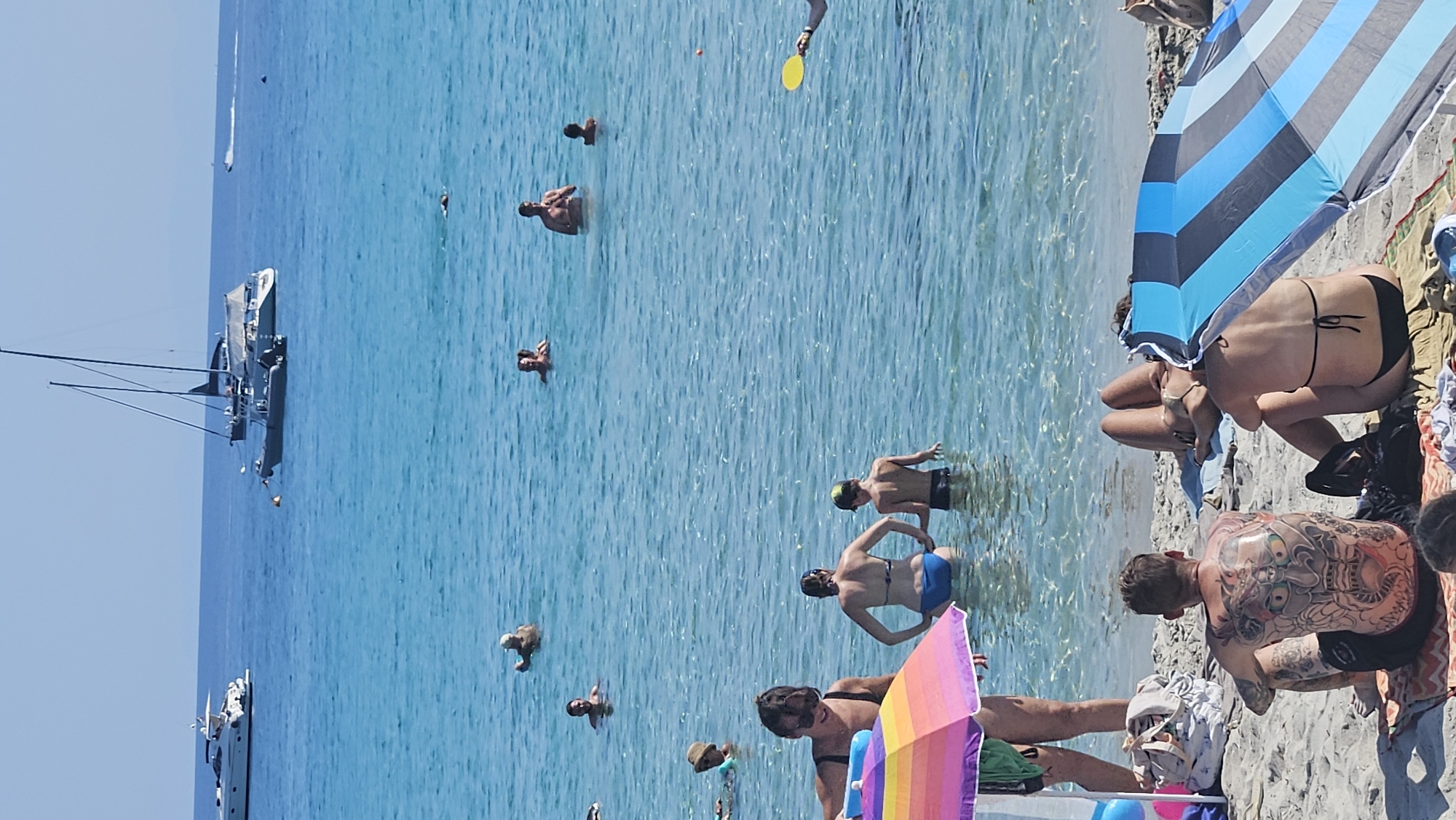 The image depicts a sunny day at a beach in L'Île-Rousse, France. The foreground shows a sandy beach with several people relaxing. A man with extensive back tattoos is seated on a towel, facing away from the camera. To his right, a woman in a black bikini is also seated, partially obscured by a blue and black striped umbrella. Further back, a woman in a bikini lies on a towel.
The middle ground features clear, turquoise water where various individuals are swimming and wading. Two adults and a child are in the shallow water, with the adults in swimsuits. More people are visible further out in the water, some appearing to be socializing.
In the background, several boats are anchored in the bay. These include a large white yacht on the left, a sailboat with two masts, and a smaller speed boat. The sea is calm and the sky is a clear blue. The overall scene conveys a relaxed atmosphere of a summer beach day.
