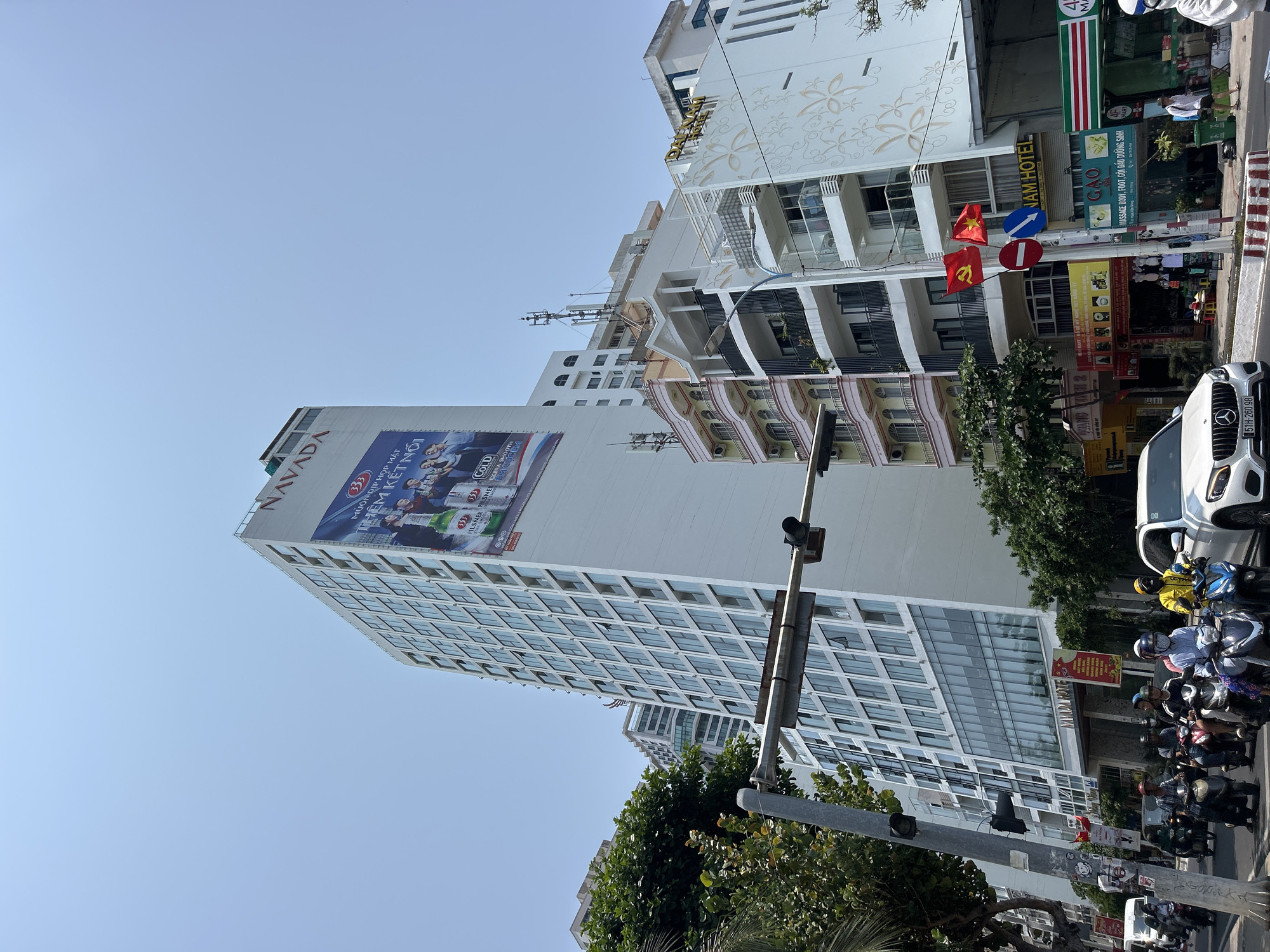 The image shows a street scene in Bắc Nha Trang, Vietnam. A tall, white building with numerous windows dominates the left side of the frame; the "NAVADA" logo is prominently displayed near the top of the building, along with a large advertisement featuring people and the number "333." To the right of this building is a smaller, white building with floral patterns, identified as the "NAM HOTEL." A street sign is visible, marked "GẠO" and offering services like massage and hair treatment. The foreground is filled with parked motorbikes and a white car. Vietnamese flags are seen, adding a national context to the setting. A traffic light pole cuts across the frame. The sky is a clear, light blue, indicating a sunny day.