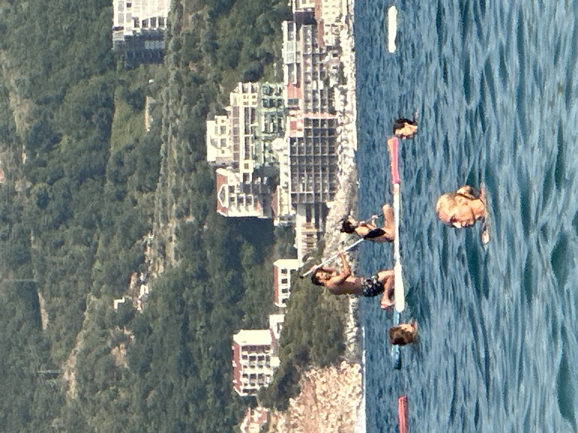 In the city of Boreti, Montenegro, a group of individuals are engaged in water activities. The scene is set against a backdrop of lush green hills dotted with buildings and residential complexes. In the foreground, several people are in the water. One woman with blonde hair is swimming, and another person's head is visible further back. In the middle ground, two people are on a stand-up paddleboard. A man, shirtless and wearing patterned shorts, is kneeling and holding a paddle. Beside him, a woman in a black swimsuit is also on the paddleboard. A young child is visible in the water to the left of the paddleboarders. Other inflatable items, likely for flotation, are present in the water. The water is a deep blue, with visible ripples indicating gentle movement. The buildings in the background appear to be hotels or apartments, suggesting a coastal resort area. The overall atmosphere is one of leisure and recreation on a sunny day.