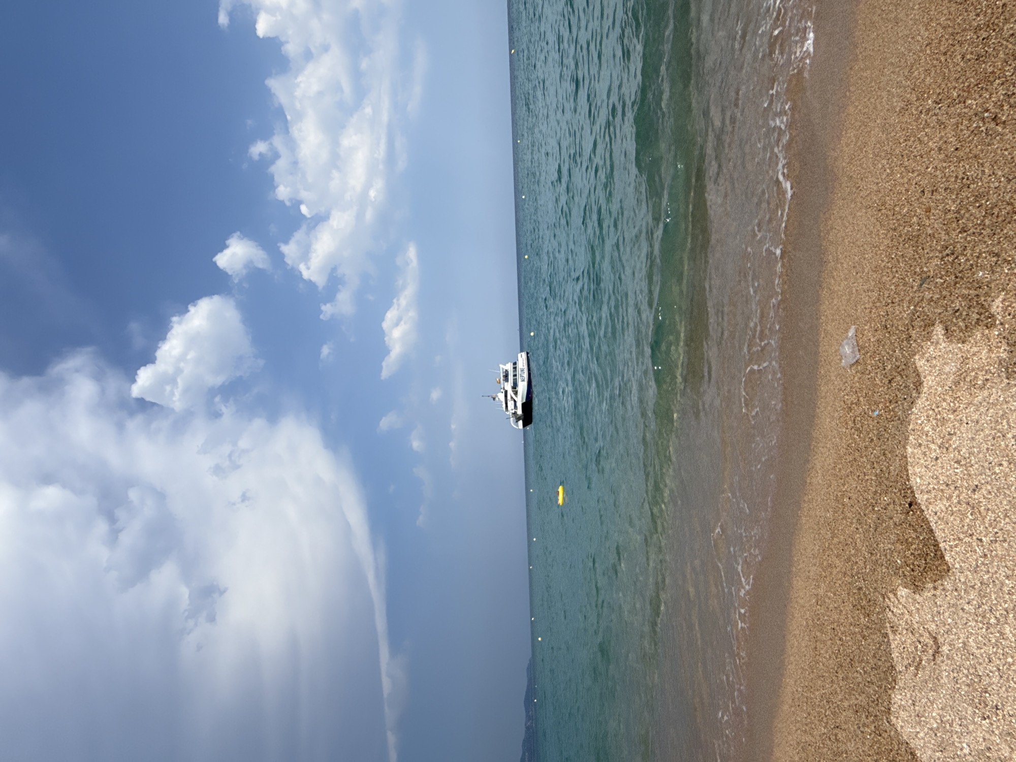 A boat with "NEPTUNE" visible on its side is in the water off a beach. The boat is white with black accents. It is a sunny day with a few clouds in the blue sky. The water is a clear, greenish-blue. There are several yellow buoys in the water. The beach is made of sand and small pebbles. There is a bit of plastic trash on the beach. The image appears to be taken from ground level, looking out at the water and sky. The sun is high in the sky, suggesting it is midday.