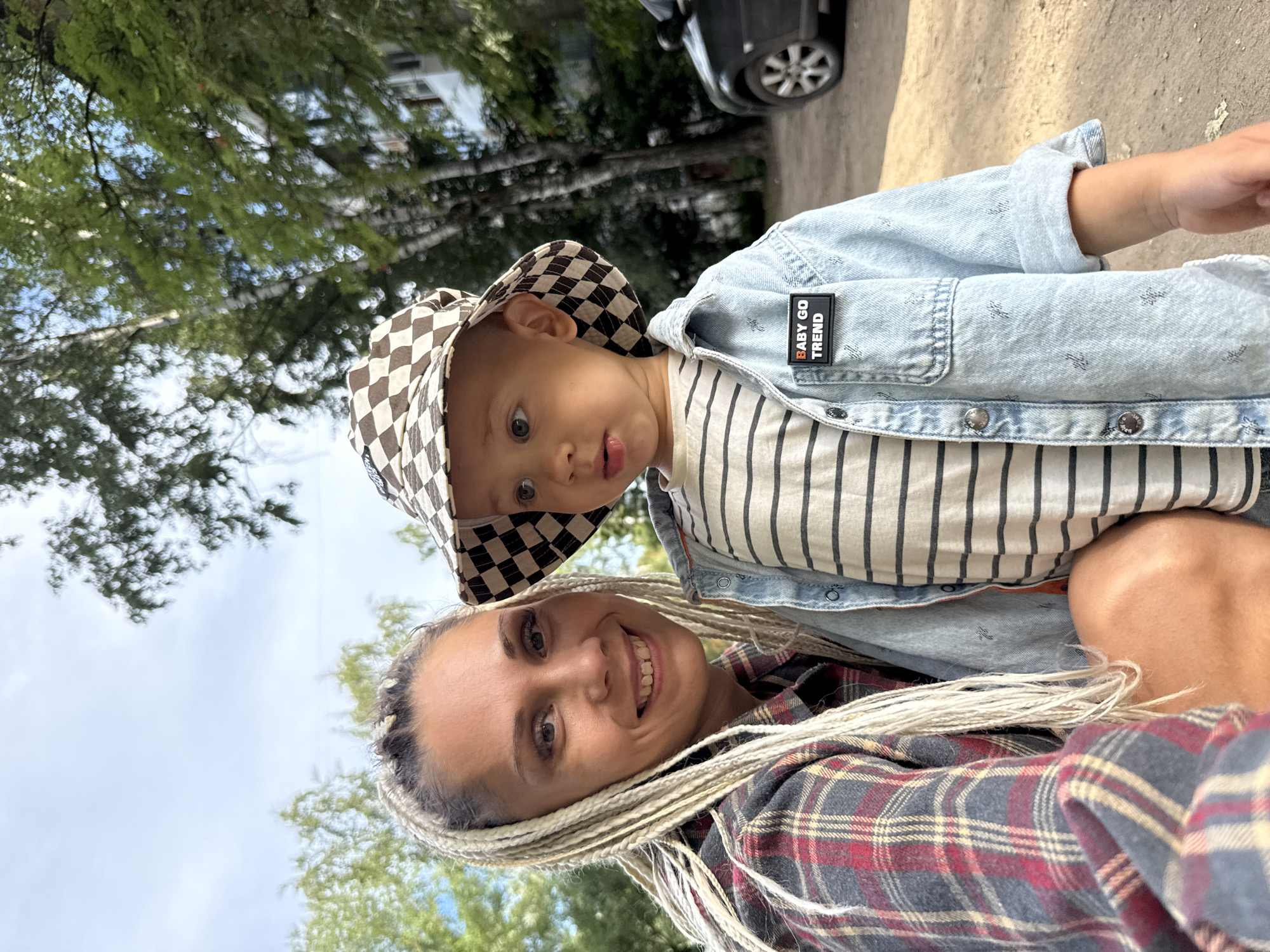 In Rusanovo, Russia, on a sunny day, a woman with blonde braided hair and a red and grey plaid shirt smiles at the camera, holding a young child. The child, wearing a checkered bucket hat and a light blue denim jacket over a striped shirt, looks to the left. The background shows trees, a building, and a parked car.