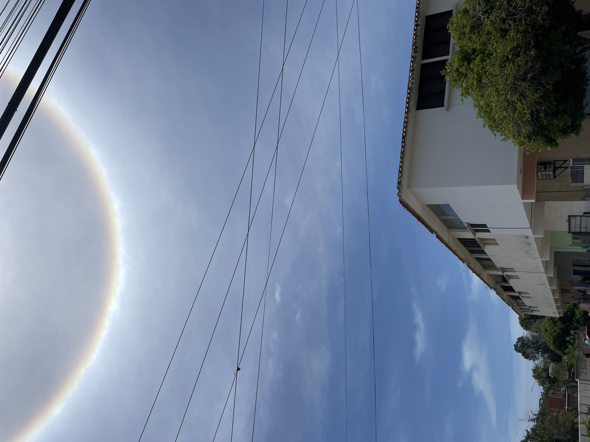 The image shows a view looking up at the sky in Porlamar, Venezuela. A bright halo surrounds the sun, which is visible in the left side of the frame. Power lines crisscross the sky. A white building with multiple stories and some visible windows is on the right. A tree is next to the building.