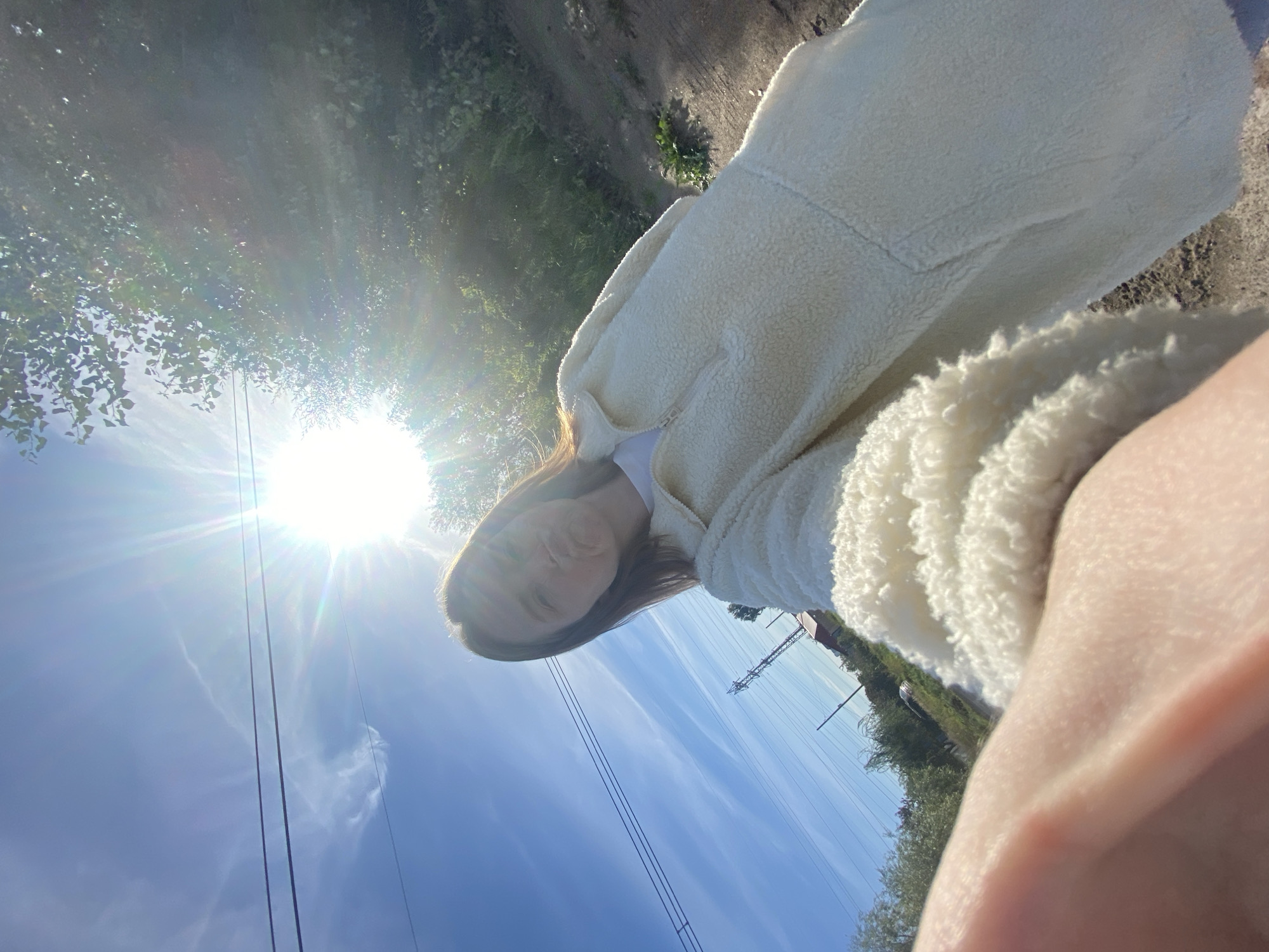 A young woman with shoulder-length brown hair is taking a selfie outdoors on a sunny day. She is wearing a fluffy, cream-colored quarter-zip jacket over a white t-shirt and is making a kissing face at the camera. Her right hand is visible in the foreground, partially obscuring the lower left corner of the frame. In the background, a clear blue sky with wispy clouds is visible, with two power lines running horizontally across it. A bright sun is positioned in the upper right quadrant, causing lens flare and washing out some of the surrounding greenery. Trees and foliage line the right side of the image, and a dirt path or road is visible on the ground. A utility pole with crossbars and insulators is located in the middle ground, to the left of the woman. A distant building with a red roof can be seen behind the utility pole. The scene appears to be located in a rural or semi-rural area, possibly on the outskirts of Chaikovsky, Russia, given the presence of utility infrastructure and natural elements.
