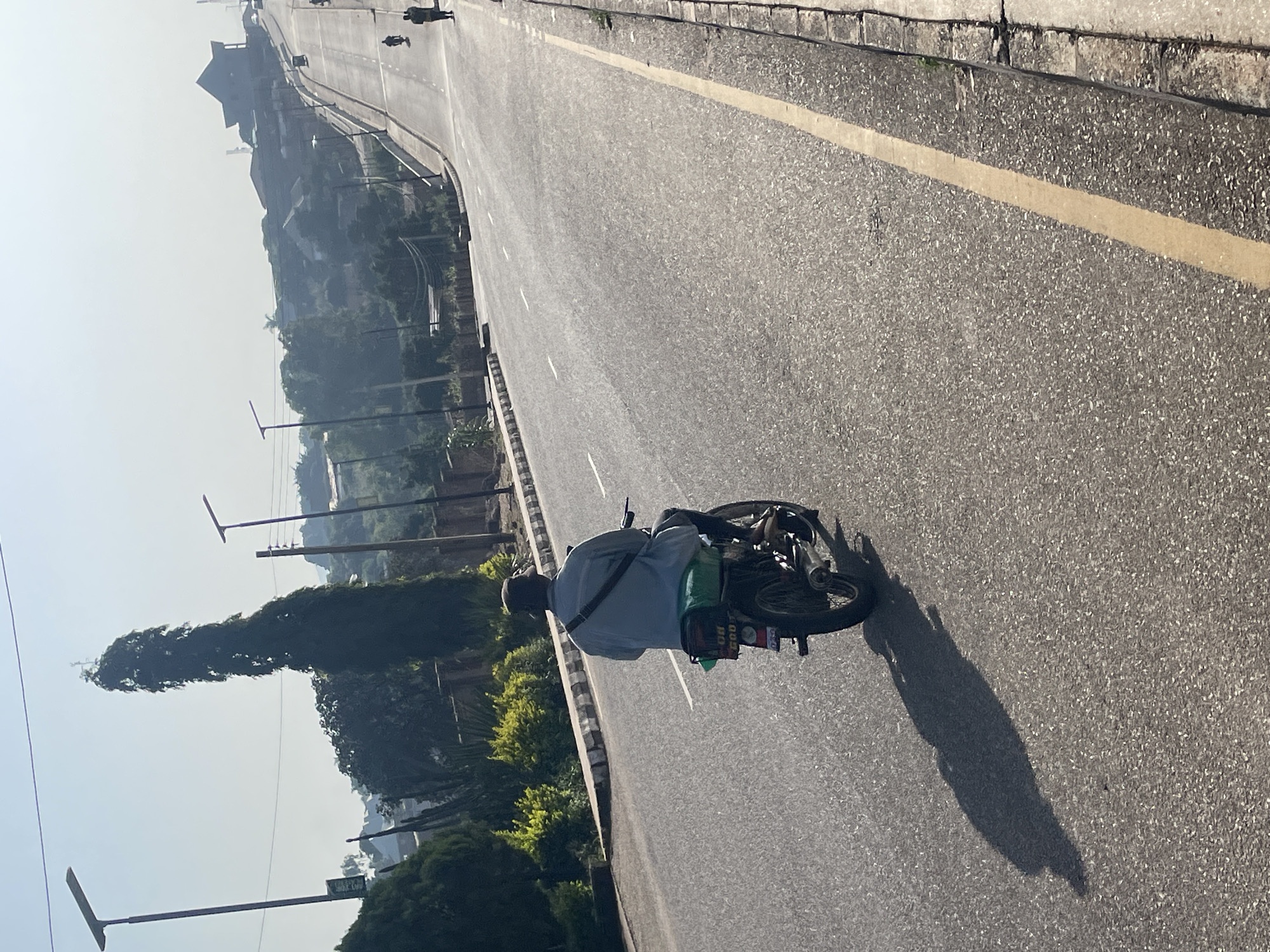 A lone motorcycle rider, wearing a blue long-sleeved shirt, dark pants, and a hat, is traveling away from the viewer on a paved road. The rider is seated on a dark-colored motorcycle with a green seat cover and a license plate that reads "GH 680 D". The motorcycle is casting a long shadow on the asphalt road. The road is an uphill incline, with a yellow line marking the edge of the lane. On the left side of the road, there are trees and bushes, as well as some residential buildings and structures in the distance. On the right side of the road, there is a concrete sidewalk or curb. The sky is clear and bright, suggesting it is daytime. The scene appears to be in a city, possibly Jos, Nigeria, given the architectural styles and vegetation visible in the background.