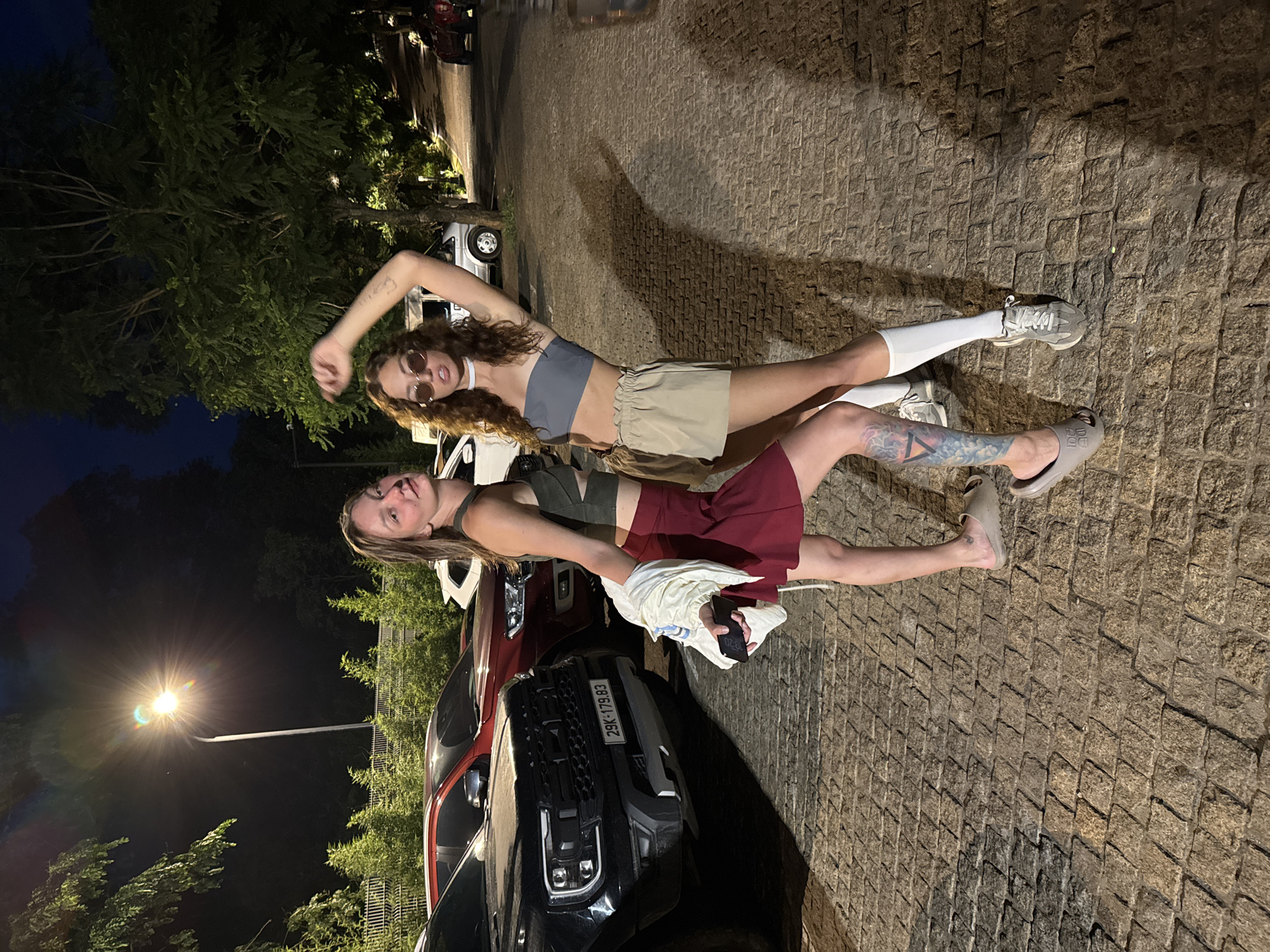 Two young women are posing for a photograph outdoors at night in Púng Luông, Vietnam. The scene is illuminated by artificial light, including a bright streetlight visible in the background and vehicle headlights. The ground is paved with cobblestones.
On the left, a woman with curly, auburn hair is wearing a grey bandeau crop top, khaki shorts, white knee-high socks, and light-colored sneakers. She has round sunglasses on and her right arm is raised above her head, with her hand open. Her expression is relaxed as she looks towards the camera.
Next to her, on the right, a woman with straight, lighter hair is dressed in a dark olive-green crop top and a maroon mini skirt. A light-colored jacket or sweater is draped over her left arm, and she is holding a dark-colored smartphone that displays "10:20". She is wearing dark slides and has a visible tattoo featuring a geometric design on her lower left leg. She is also looking towards the camera with a calm expression.
Behind the women, several vehicles are parked, including a white sedan and a dark-colored SUV or pickup truck. The license plate "29K-179.83" is visible on the front of the dark vehicle. Trees and foliage are present in the background, further indicating an outdoor setting. The overall mood is casual and friendly, capturing a nighttime moment.