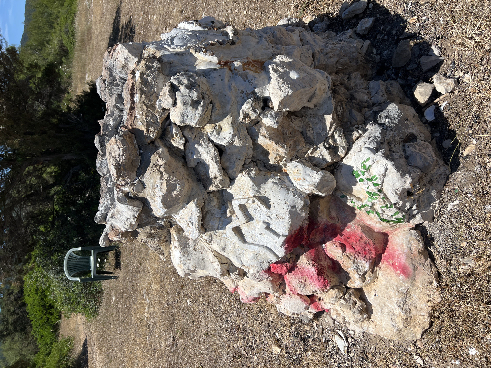 The image depicts an outdoor scene with a prominent pile of rocks in the foreground. The rocks are of various sizes and textures, with some appearing weathered and others rough. Several rocks are painted with markings. A section of the pile on the left side is adorned with bright pink paint, partially obscuring the natural rock color. Above the pink paint, a symbol resembling a stylized 'M' or a branched design is carved or painted in a light color, possibly white, onto a larger rock. To the right of this symbol, a small cluster of green painted elements, resembling leaves or vines, is visible on another rock. The ground surrounding the rock pile is covered in dry, sparse vegetation and scattered stones.
In the background, a green plastic chair is positioned amongst dense green bushes and under the shade of a tree. Further back, a hilly landscape with more trees and foliage is visible under a clear blue sky. The overall environment suggests a natural, possibly rural or coastal setting. The presence of the chair and the deliberate markings on the rocks hint at human activity or a site of interest within this natural landscape. The location is identified as being in Peschici, Italy.