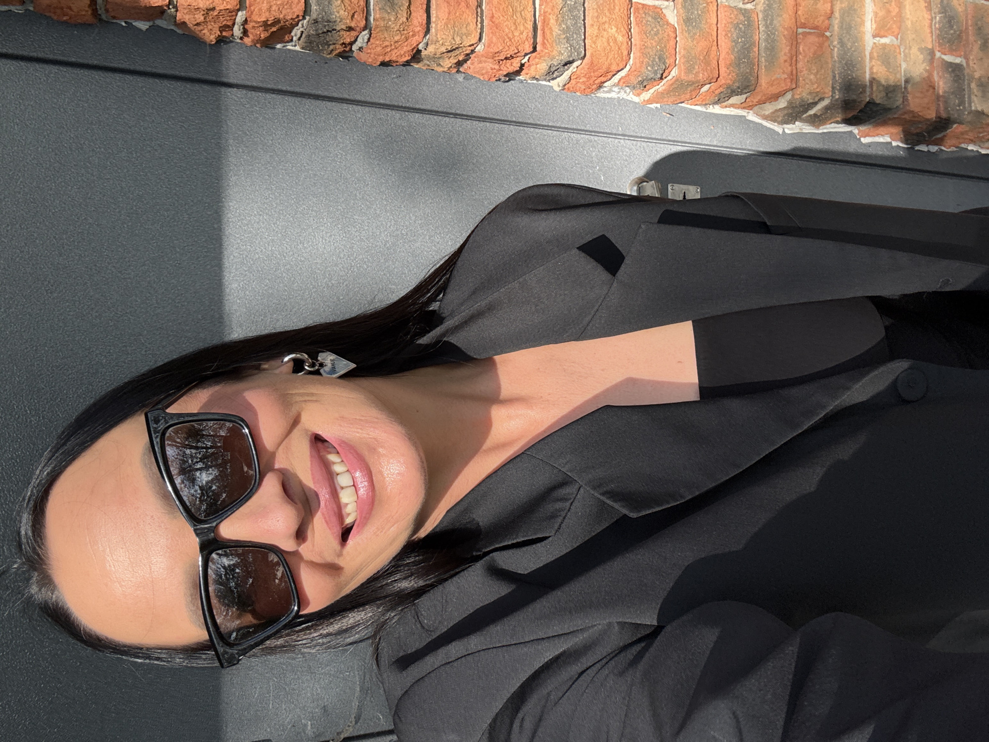 A woman with dark hair and sunglasses smiles at the camera. She is wearing a black blazer over a black top and silver heart-shaped earrings. To her right is a brick wall and to her left is a gray door. The image is taken outdoors. The location of Chaikovsky, Russia is not discernible from the image.