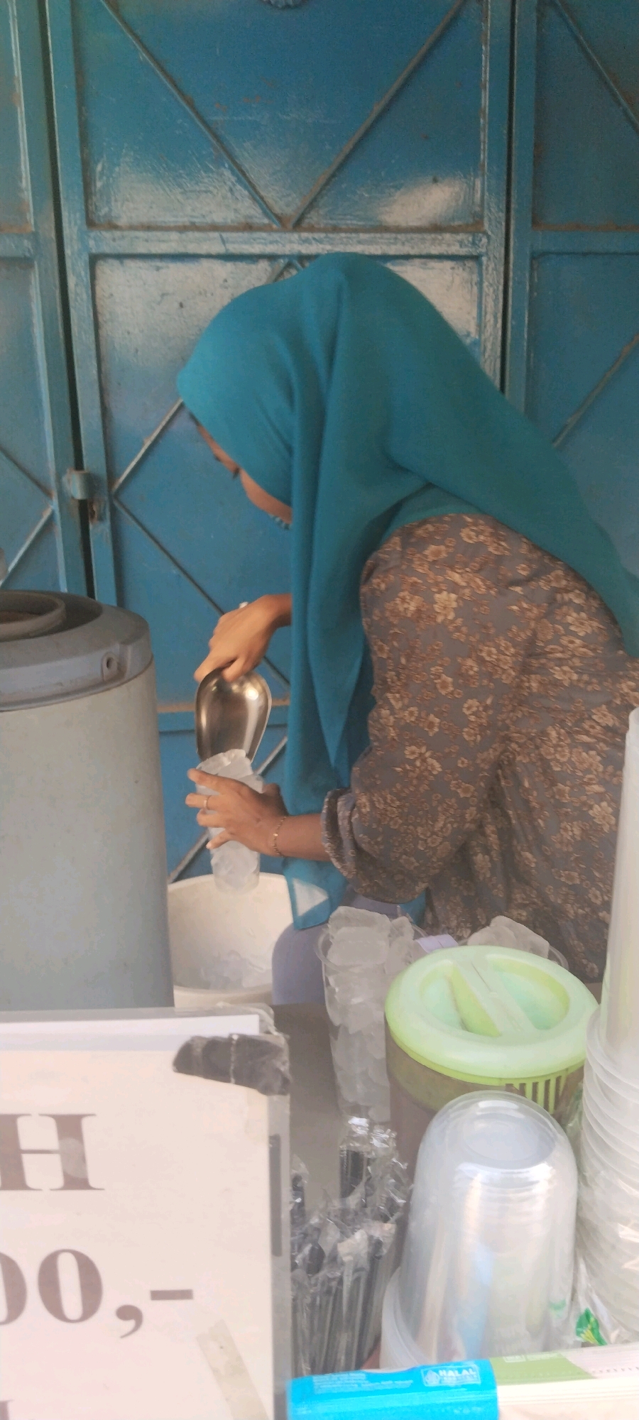In West Jakarta, Indonesia, a woman wearing a blue hijab and a floral-patterned dress is seen preparing a drink. She is scooping ice from a large container using a metal scoop and placing it into a clear plastic cup. The scene appears to be outdoors, possibly at a street food stall or a small shop, given the blue metal gate in the background and the various containers of ice and cups on a table in the foreground. There are stacks of clear plastic cups and a green lid on a container are visible. A sign with text and numbers, possibly indicating prices or product names, is also present. The overall atmosphere suggests a commercial transaction is about to take place.