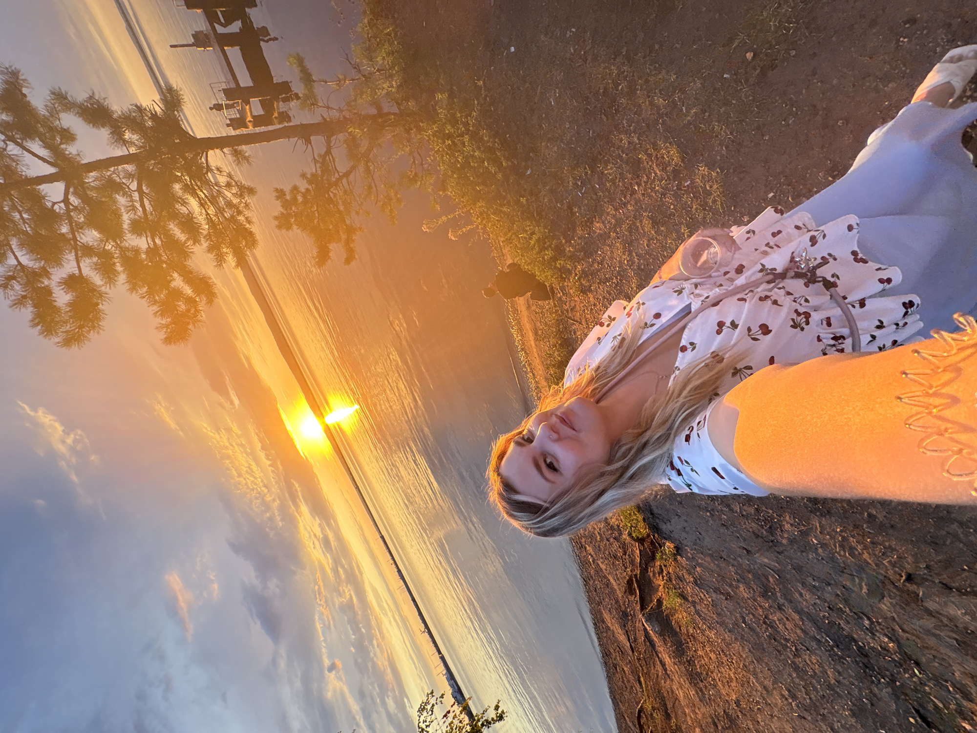 This is a selfie taken by a woman at sunset on the embankment of the Kama River in Chaikovsky, Russia. The woman has blonde hair and is wearing a white top with a cherry print and a light blue skirt. She is holding a glass of drink. In the background, the sun is setting over the river, casting a warm glow on the water and the sky. A pier with a railing and a few people can be seen on the right side of the image. A large tree is also visible on the right. The ground in the foreground is dirt and covered with some dry grass and roots.