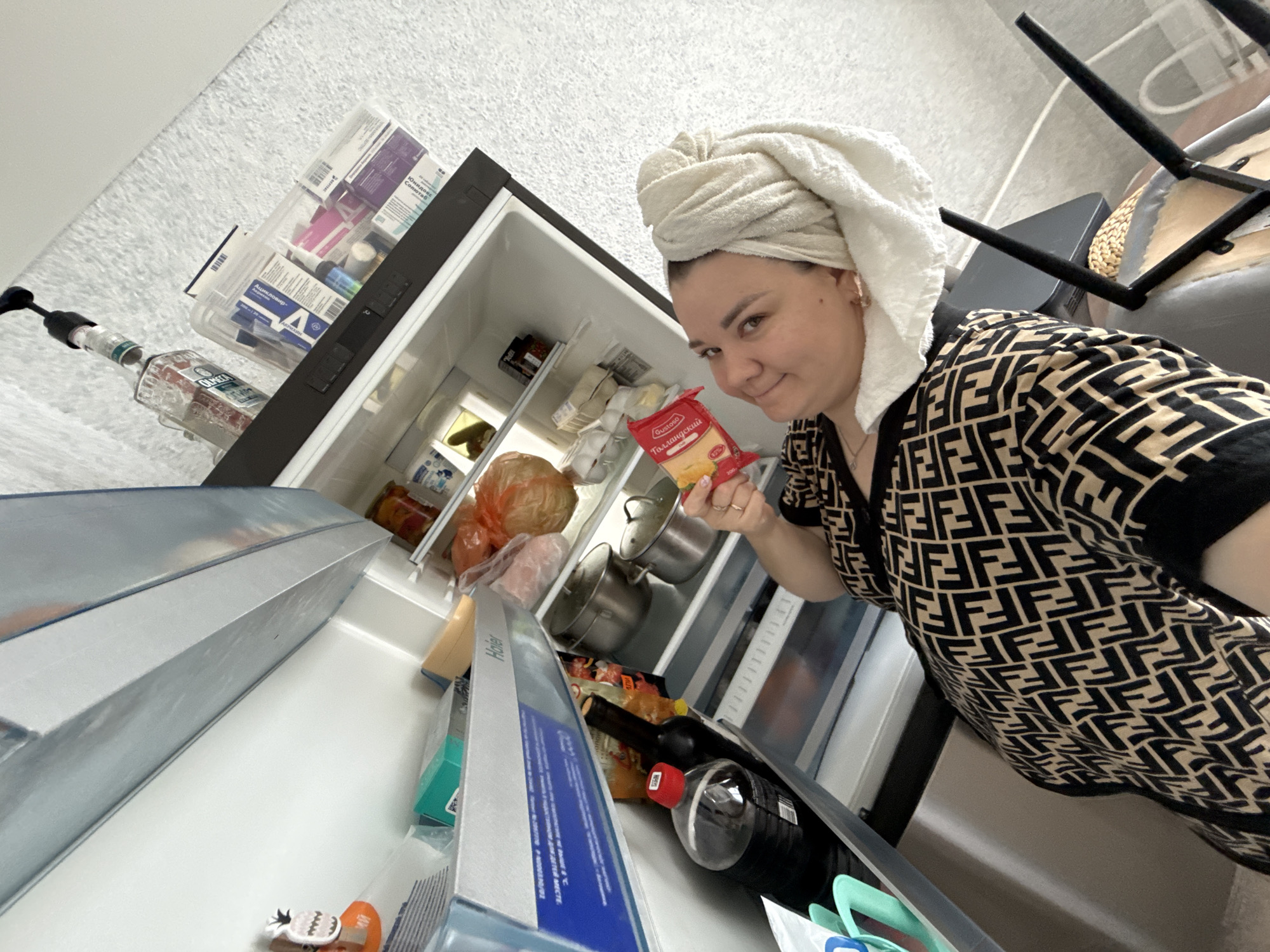 A woman with a towel wrapped around her head is standing in front of an open refrigerator. She is wearing a black and tan patterned robe and holding a package of cheese. The refrigerator contains various food items, including bottles of soda, cooking pots, and bags of produce. There are also several boxes of medication on the top shelf. The scene appears to be taking place indoors, likely in a kitchen, during the daytime. The woman is smiling at the camera, suggesting a casual and possibly humorous moment. Visible text includes brand names like "Haier," "Oro," and "Gustoso," as well as Russian text on the medication boxes.