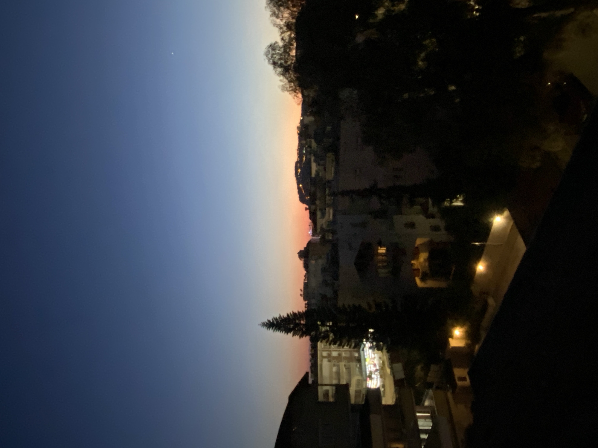 This image, captured during twilight in Alanya, Turkey, presents a panoramic view of a town from an elevated perspective. The sky exhibits a striking gradient, transitioning from a deep indigo blue at the top, gradually lightening to a soft blue-grey, and finally to a warm, pinkish-orange hue along the horizon, indicating either sunset or dawn. A single, bright point of light, likely a star or planet, is visible in the darker expanse of the sky.
Below the colorful horizon lies a sprawling townscape, with numerous buildings and structures extending across what appears to be hilly terrain. As darkness settles, artificial lights from windows and streetlamps begin to illuminate the scene, casting warm yellow and orange glows that dot the urban fabric. Several trees are visible, mostly as dark silhouettes against the fading light. A prominent, conical conifer tree stands in the middle ground, its dark foliage reaching upwards. The immediate foreground consists of dark, indistinct shapes, possibly a railing or a part of the building from which the photograph was taken, framing the expansive vista.
No specific human activity or event is depicted, focusing instead on the serene transition of day to night over the town. The weather appears clear, with no clouds visible. There is no discernible text within the image.